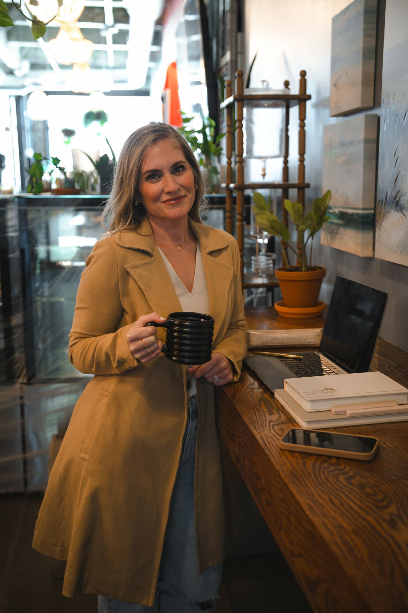 Woman with long hair in a tan coat holding a black mug, standing in a cozy cafe with plants and books.