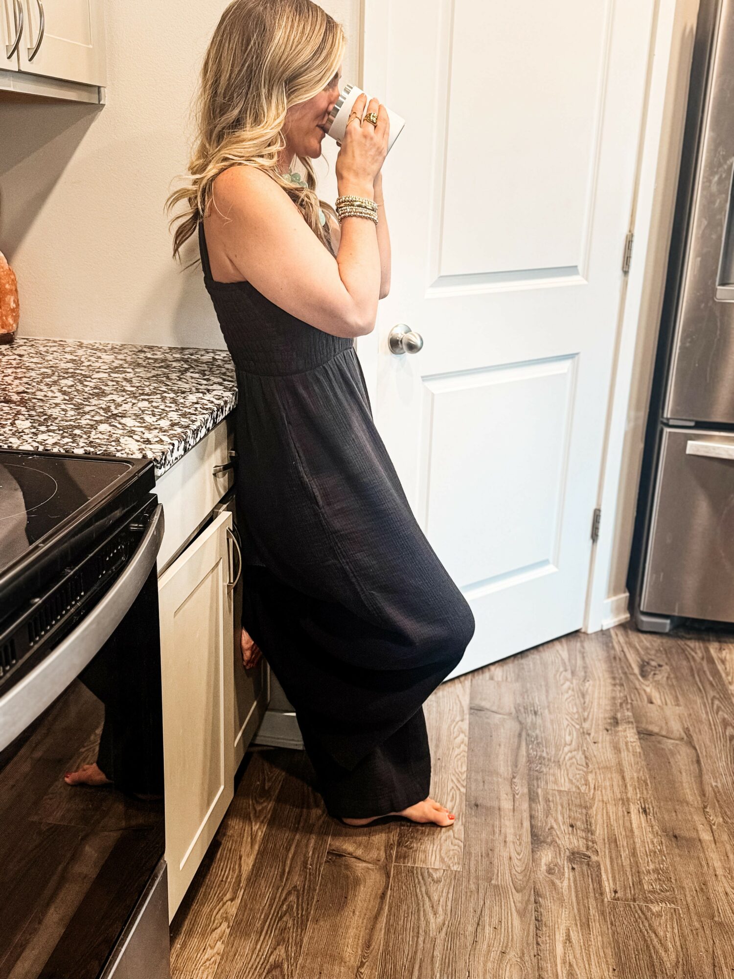 Woman with blonde hair in a black dress standing in a kitchen, leaning against a counter, holding a cup near her face.