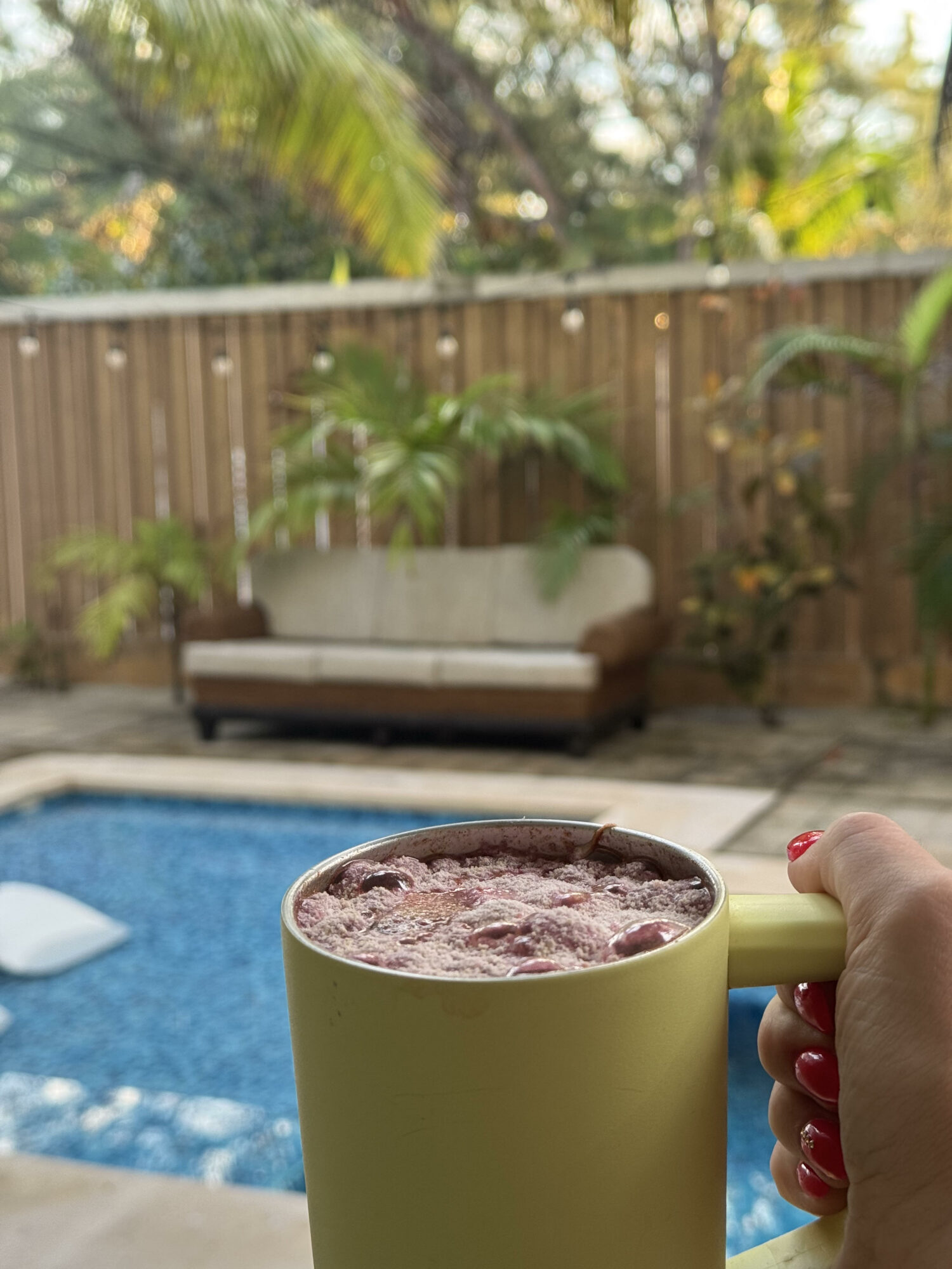 Person holding a yellow mug with a hot beverage near a swimming pool, outdoor patio with a sofa and plants in background.