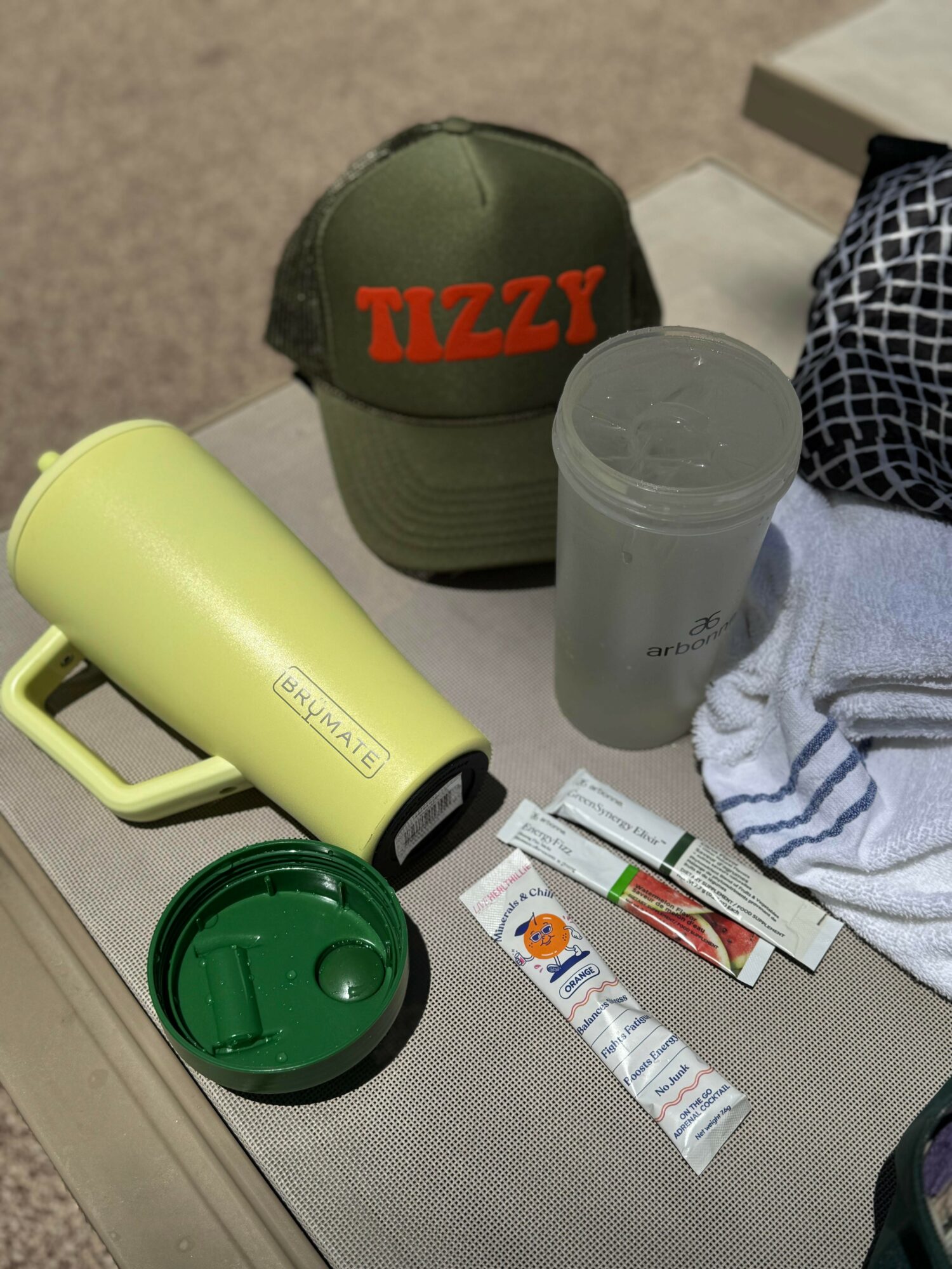 Cap with red text, water bottle, yellow mug, and snack bars on a table, with a towel nearby.