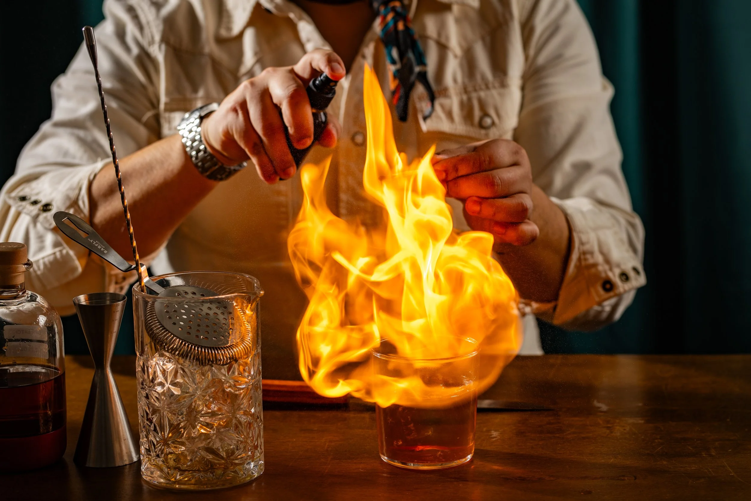 Person holding a lighter over a bowl with flames, on a wooden table, with glass and metal objects nearby.