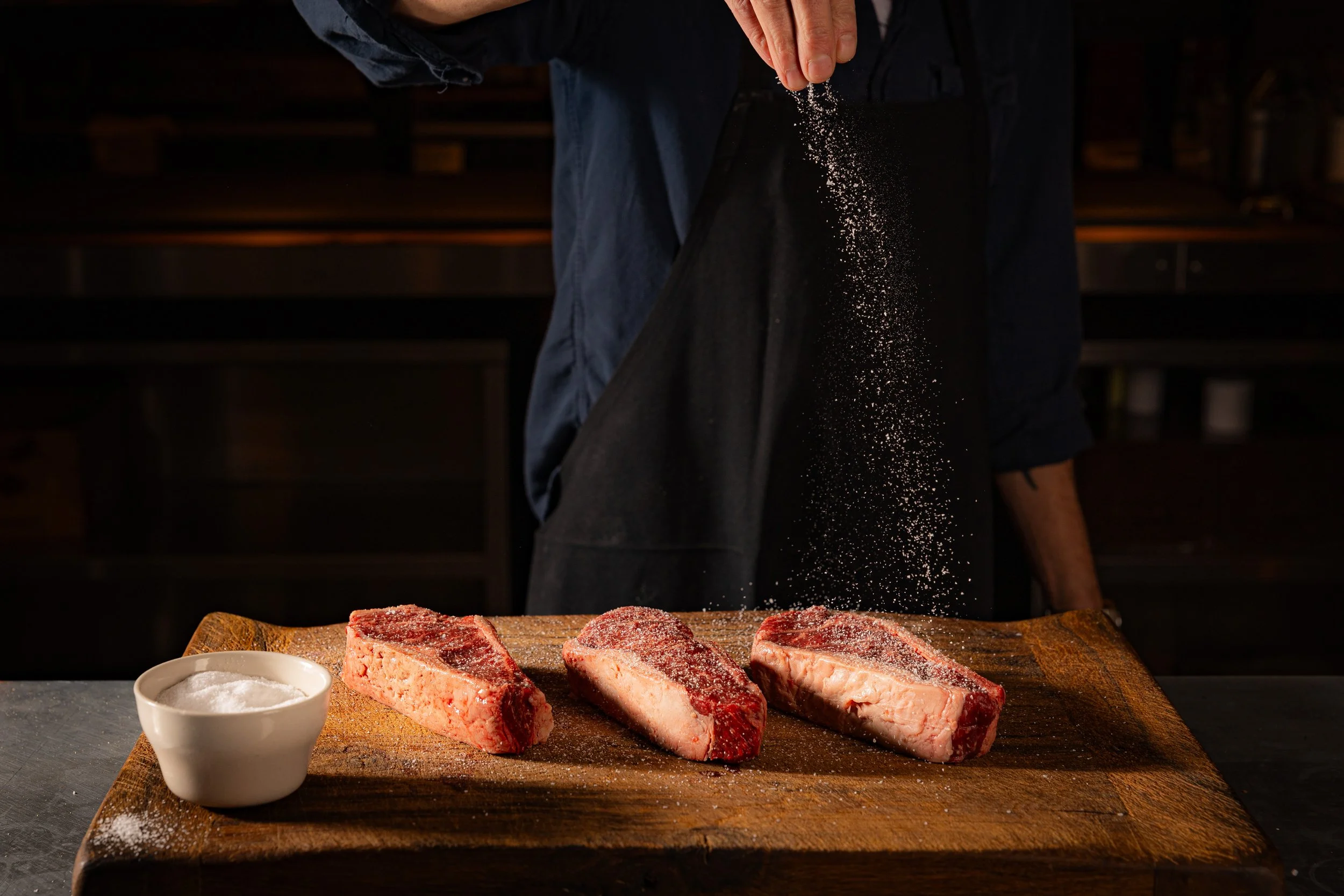 Person sprinkling salt on three pieces of raw meat on a wooden cutting board with a small bowl of salt nearby