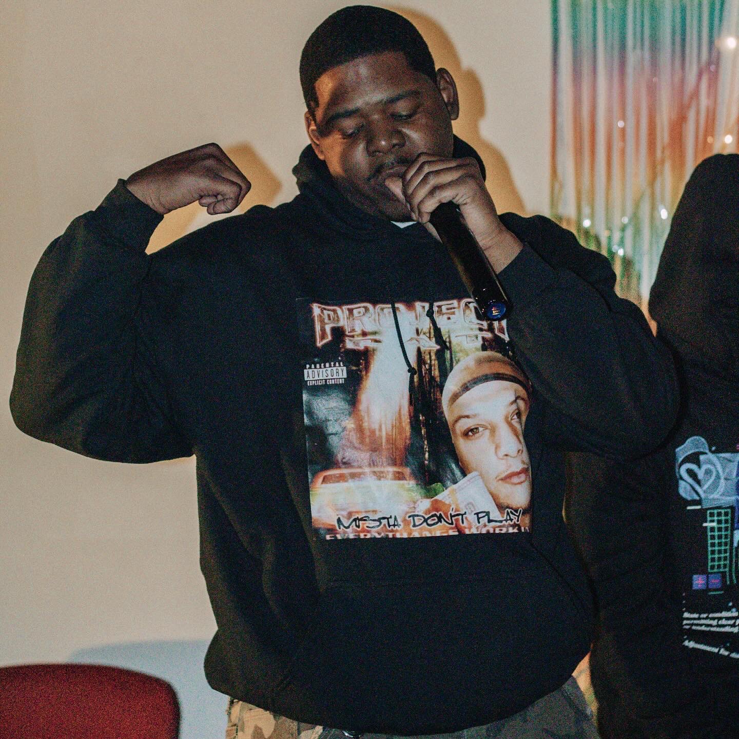 Person wearing a black hoodie with a graphic print, flexing arm, and holding a microphone, indoors with colorful curtains.