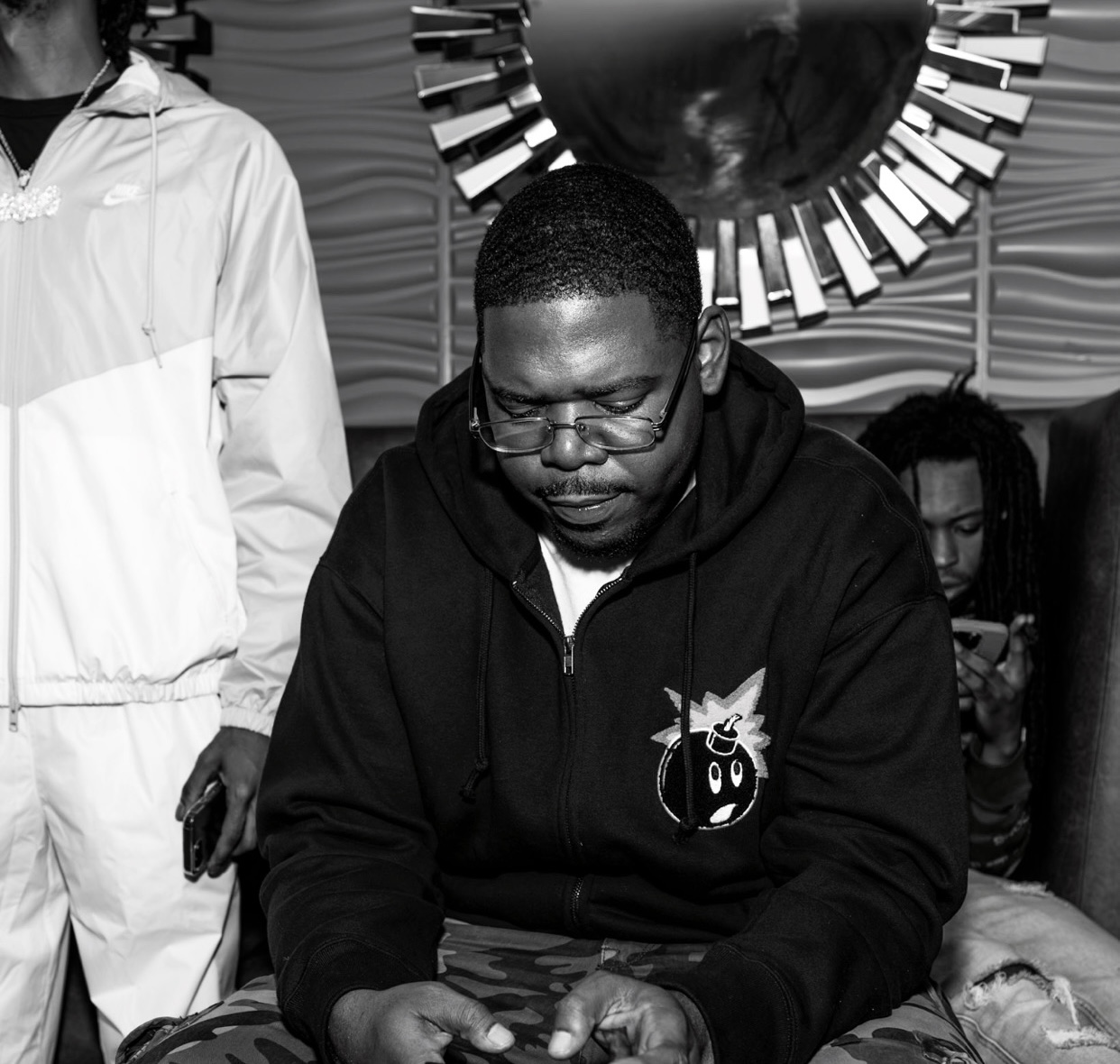 Man with glasses and a hoodie sitting, holding a phone, with a decorative mirror behind him.