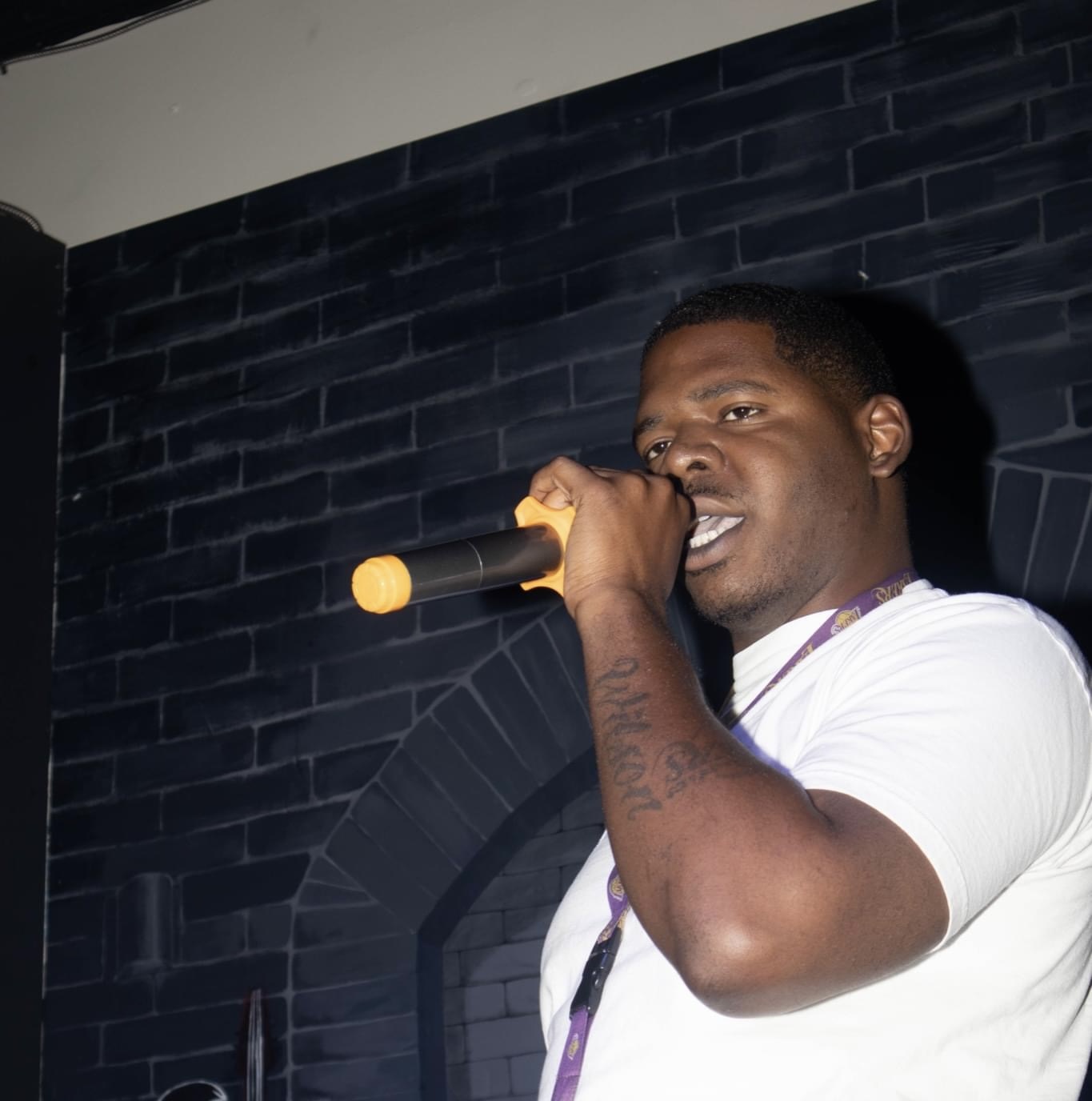 Man holding a yellow microphone, singing or speaking, against a dark brick wall background.