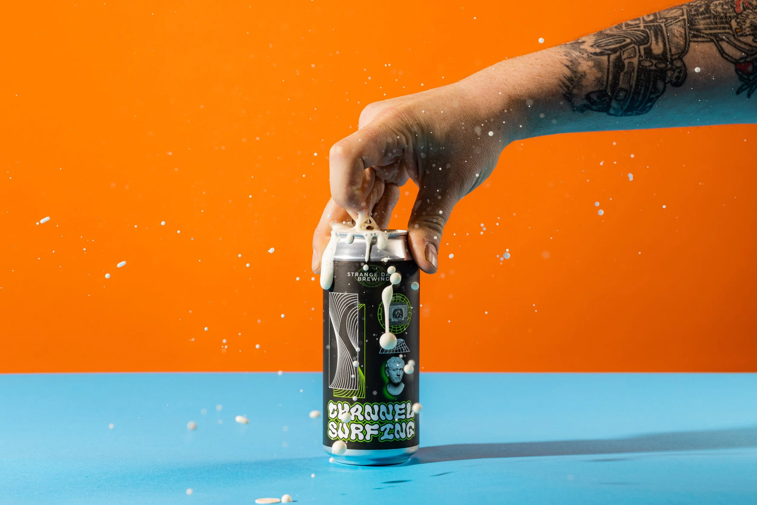 Hand with tattoo opening a can against orange background, with water splashing around, on blue surface.
