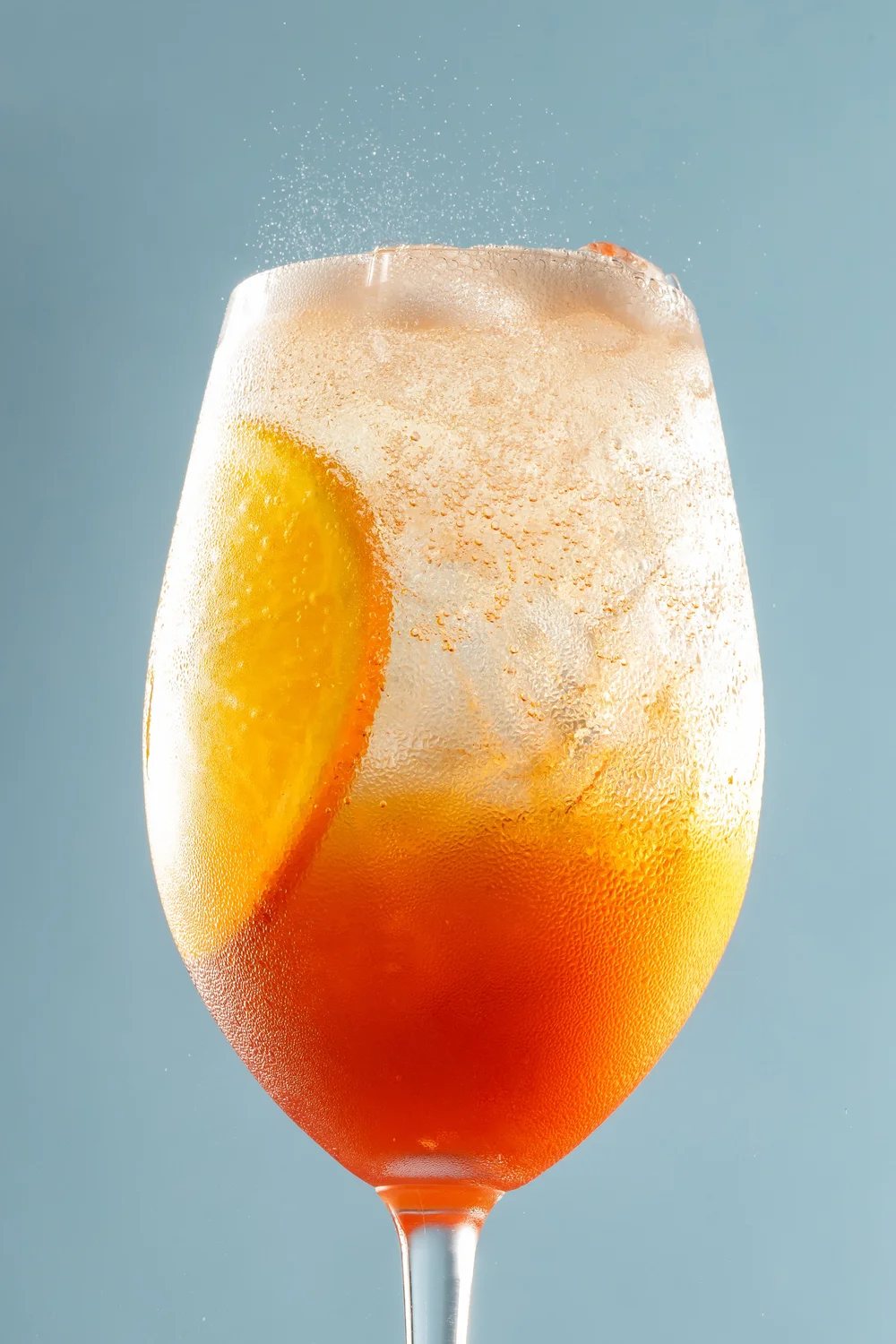 A colorful cocktail in a tall glass with a lemon wedge, topped with foam, against a light blue background.
