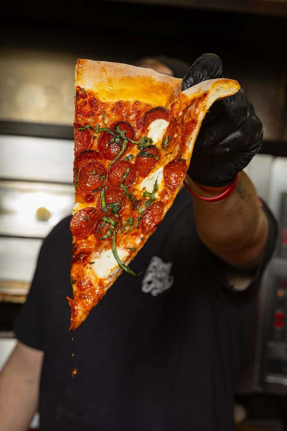 Person holding a large slice of pepperoni and green pepper pizza, wearing a black glove, in a kitchen setting.
