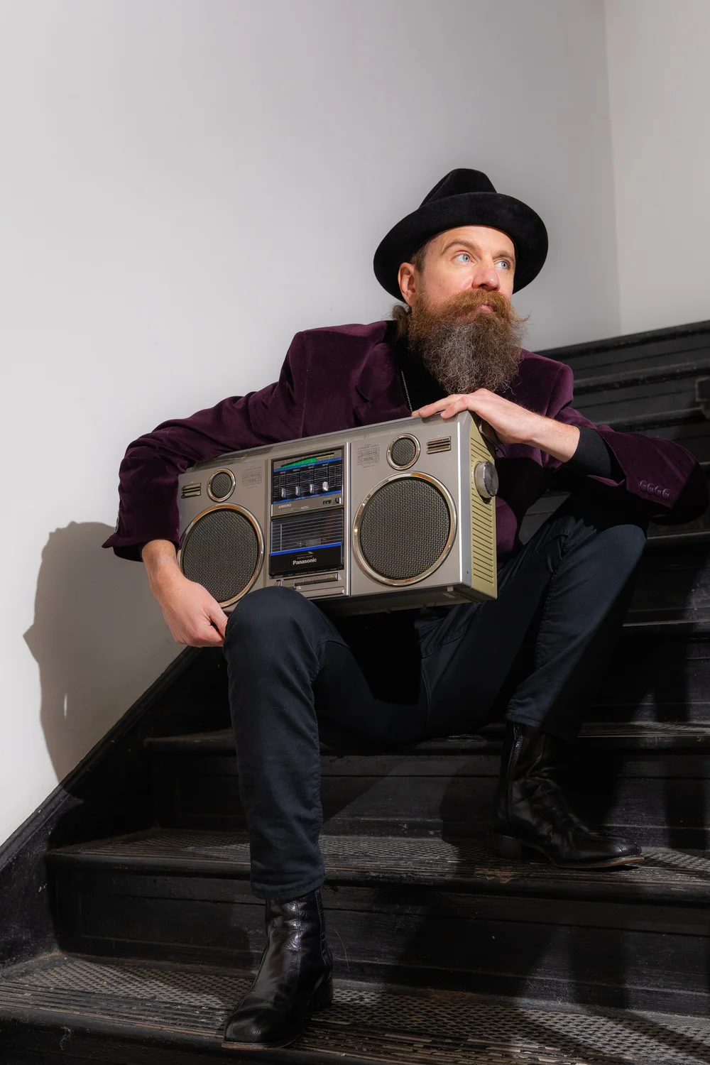 Man with a beard and hat sitting on stairs, holding a vintage boombox, looking to the side, against a plain wall.