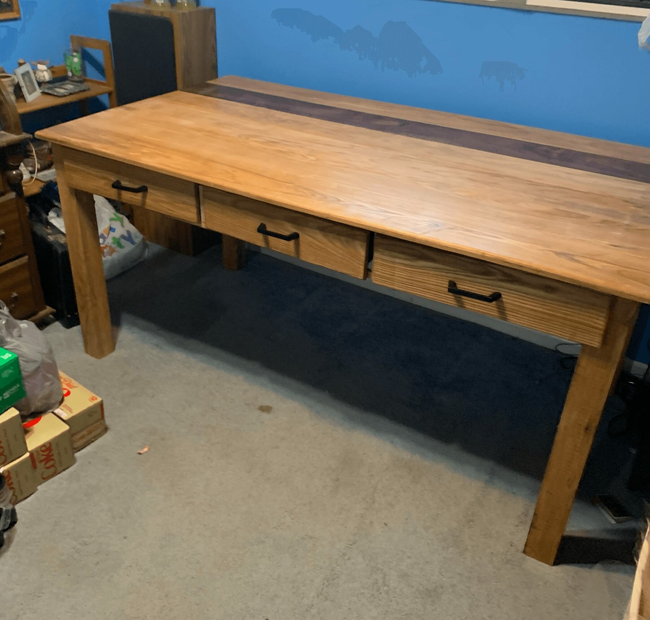 Wooden desk with three drawers and black handles, placed against a blue wall, on a carpeted floor.