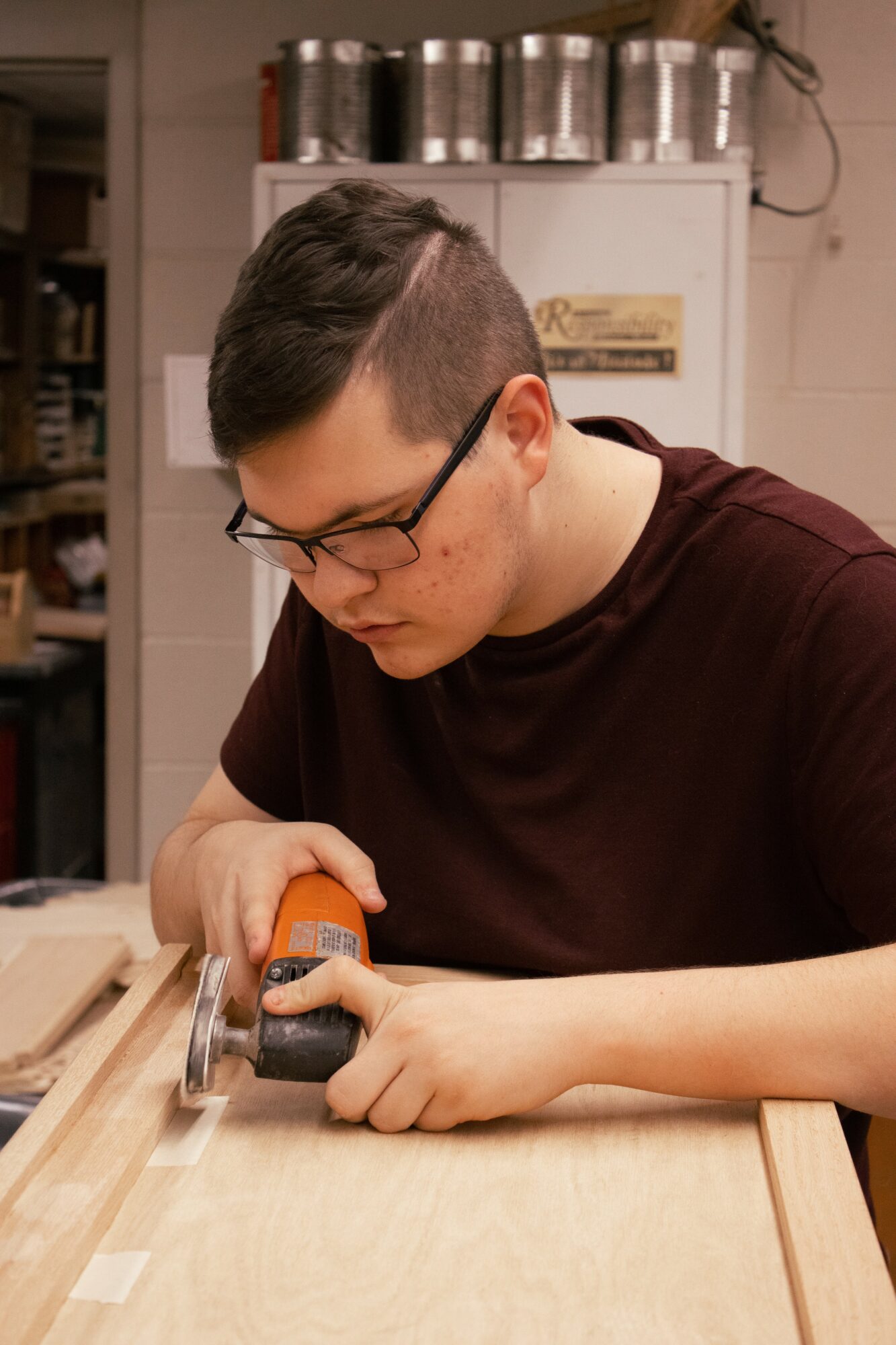 Young person with glasses using a handheld power tool on a wooden surface, focused on work, in a workshop setting.
