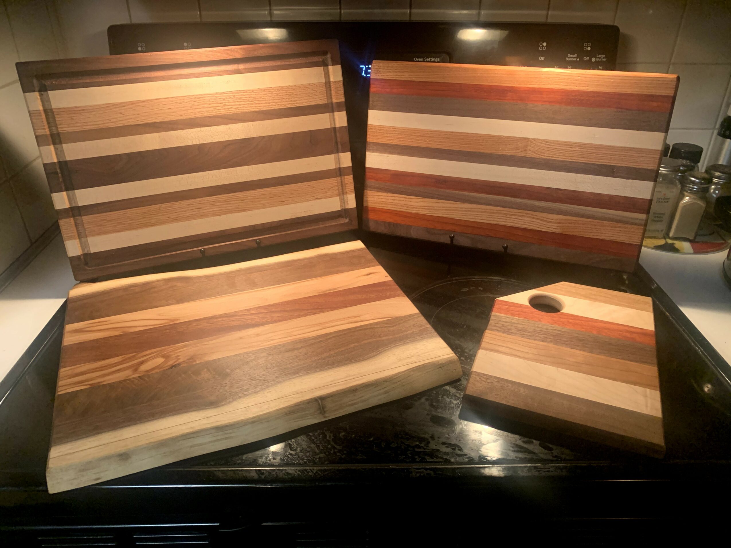 Four wooden cutting boards with striped patterns, one leaning against a wall, one flat, and one smaller with a hole, on a dark surface.