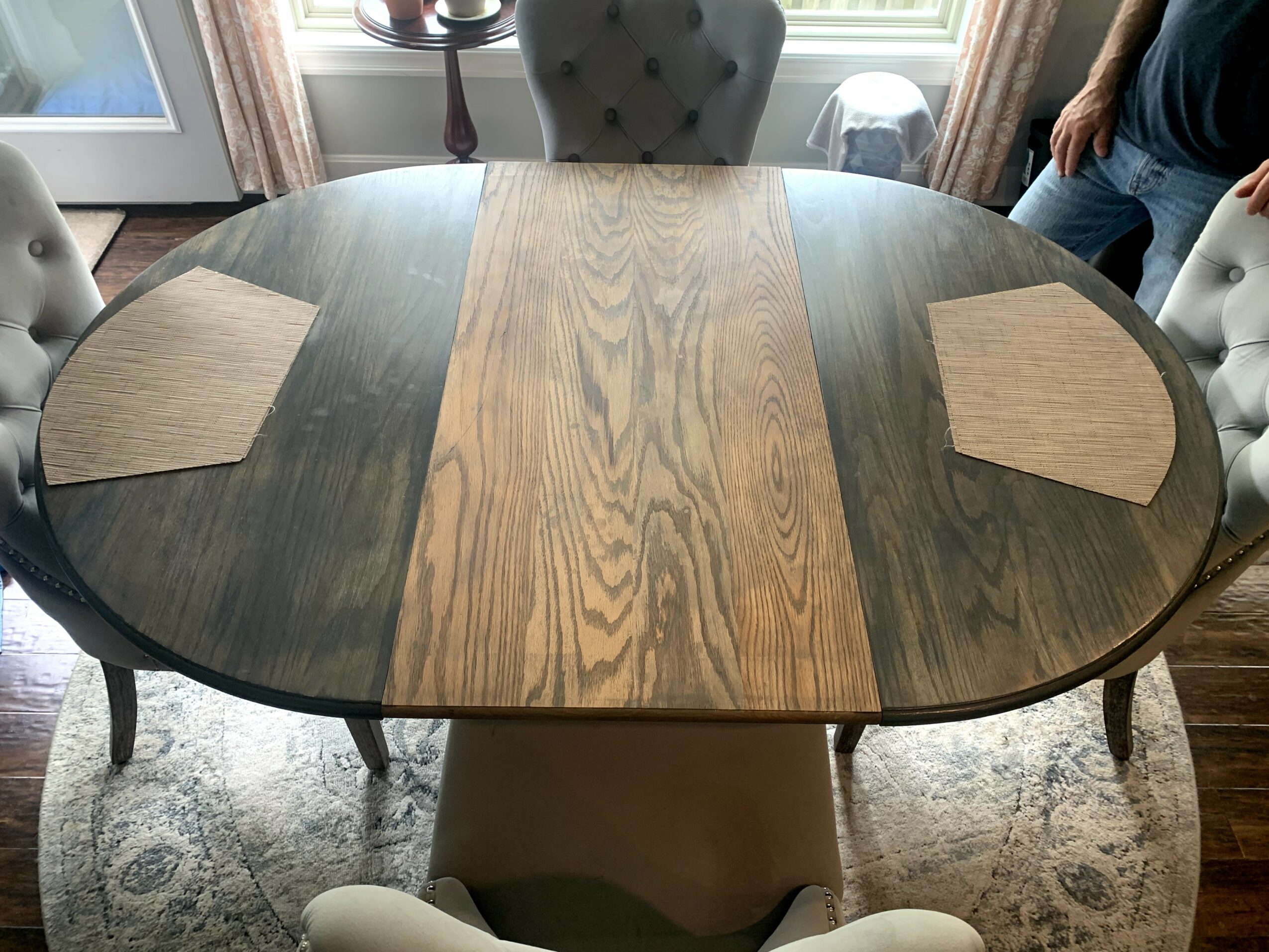 Oval dining table with a central wood panel and dark sides, surrounded by upholstered chairs, in a room with large windows.