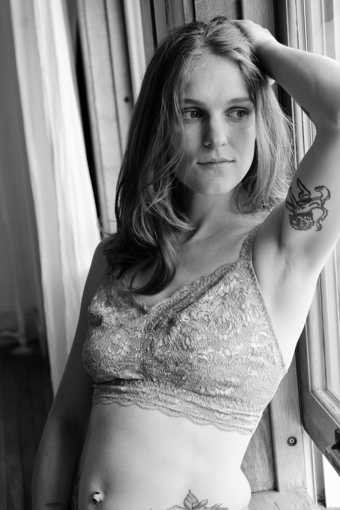 Young woman with shoulder-length hair looking out window, arm raised, tattoo on upper arm, wearing lace top, black and white photo.