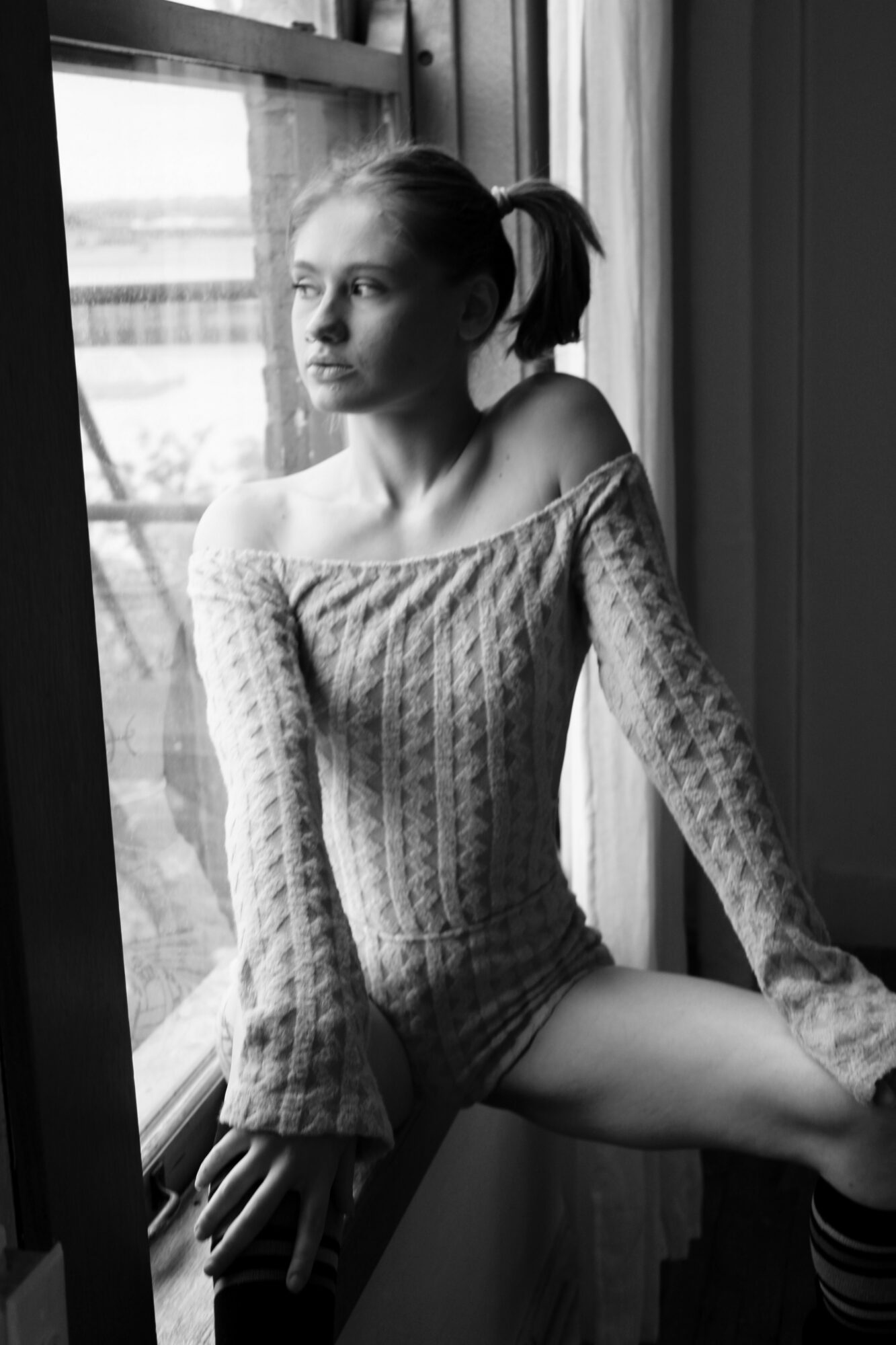 Young woman sitting by a window, looking outside, wearing a knitted sweater, with natural light illuminating her face.