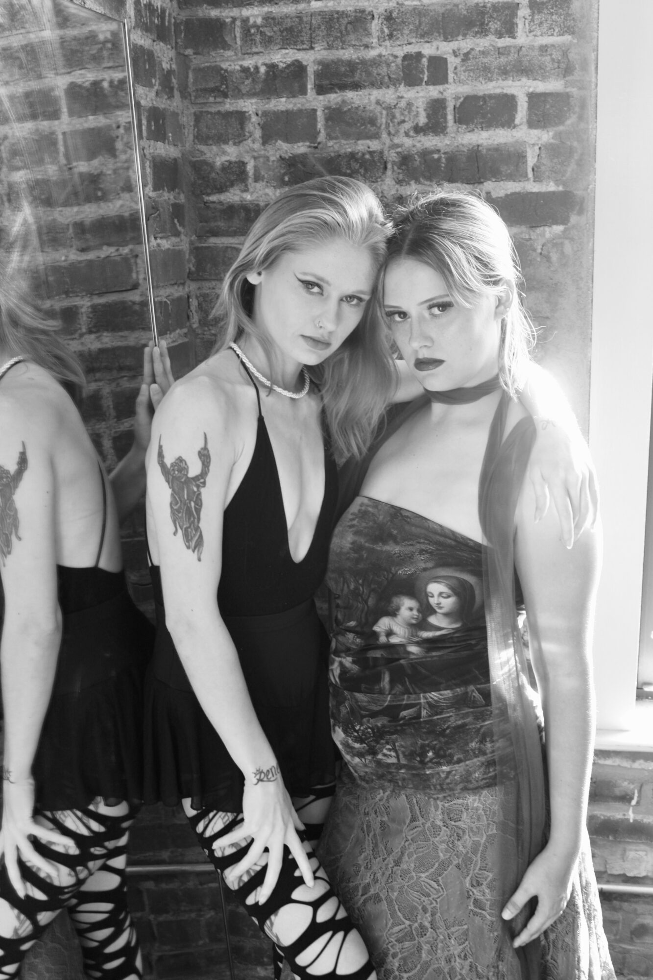 Two women with tattoos and long hair pose against a brick wall, one wearing a black dress and the other a patterned dress.