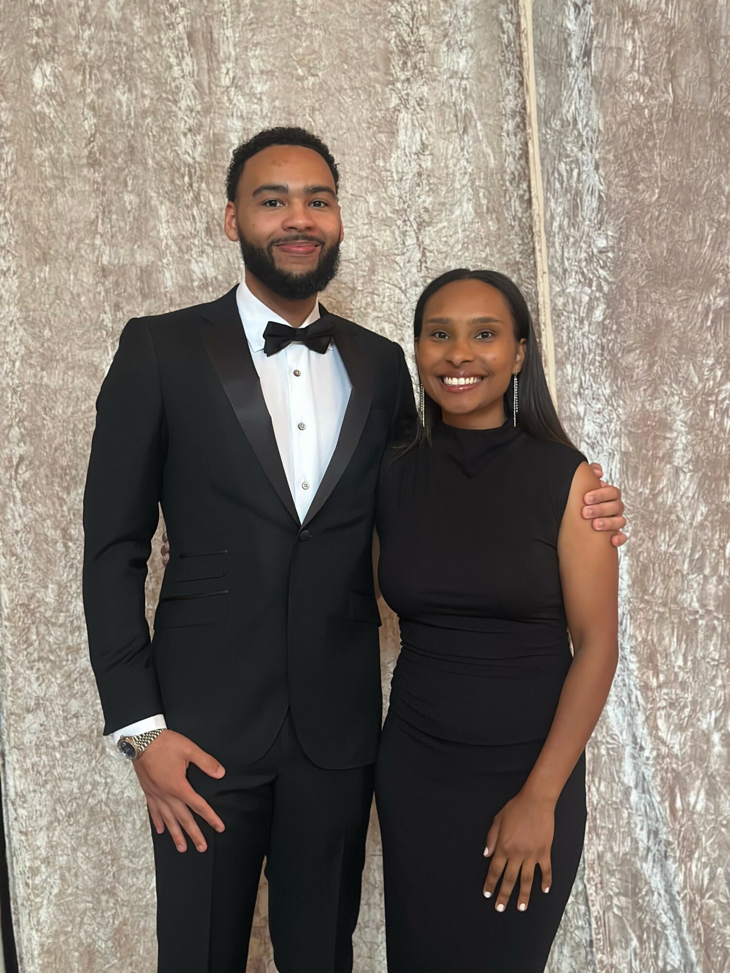 A man in a tuxedo and a woman in a black dress stand together, smiling, against a textured background.