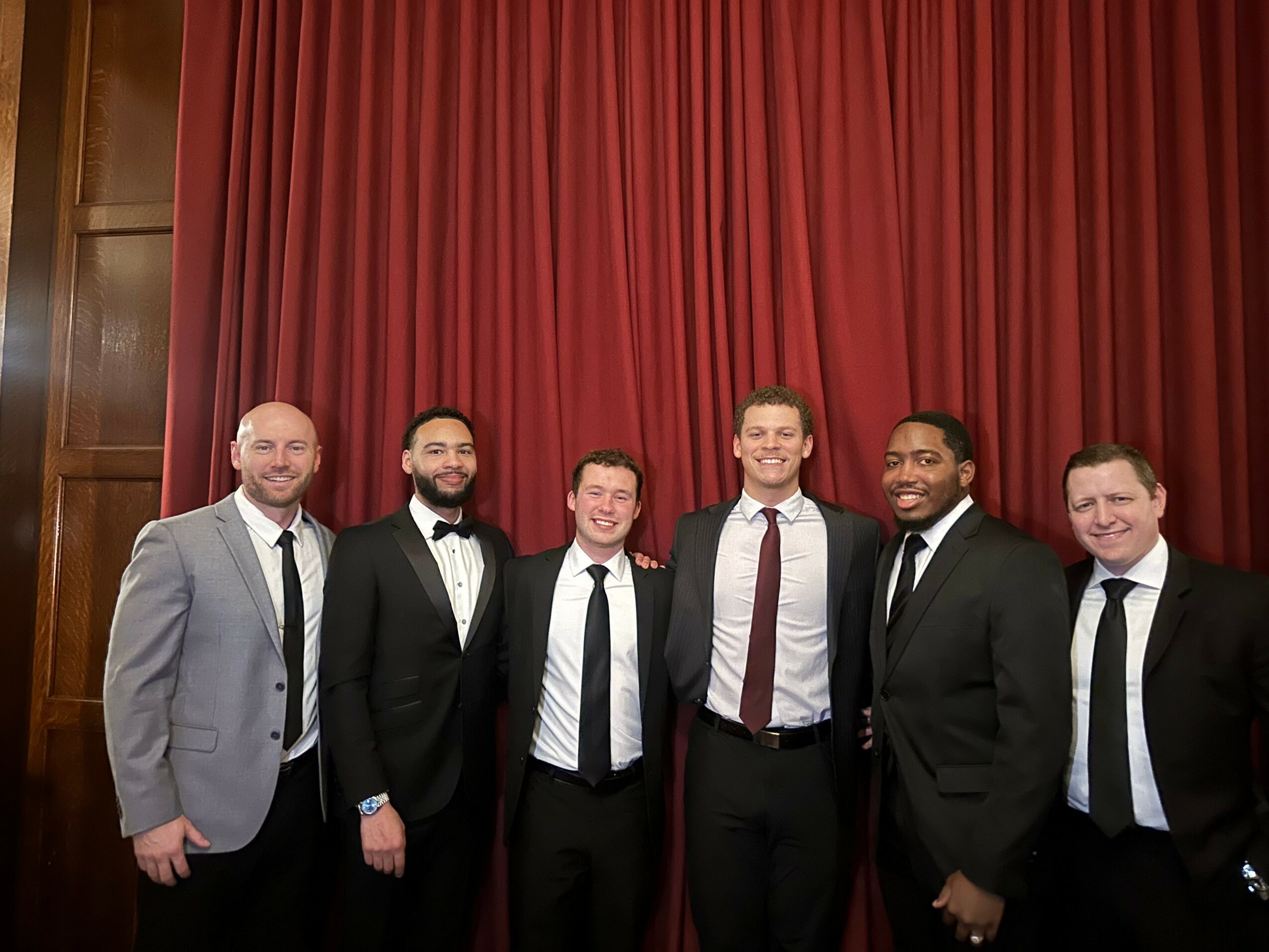 Six men in formal attire standing in front of a red curtain, smiling for a group photo.