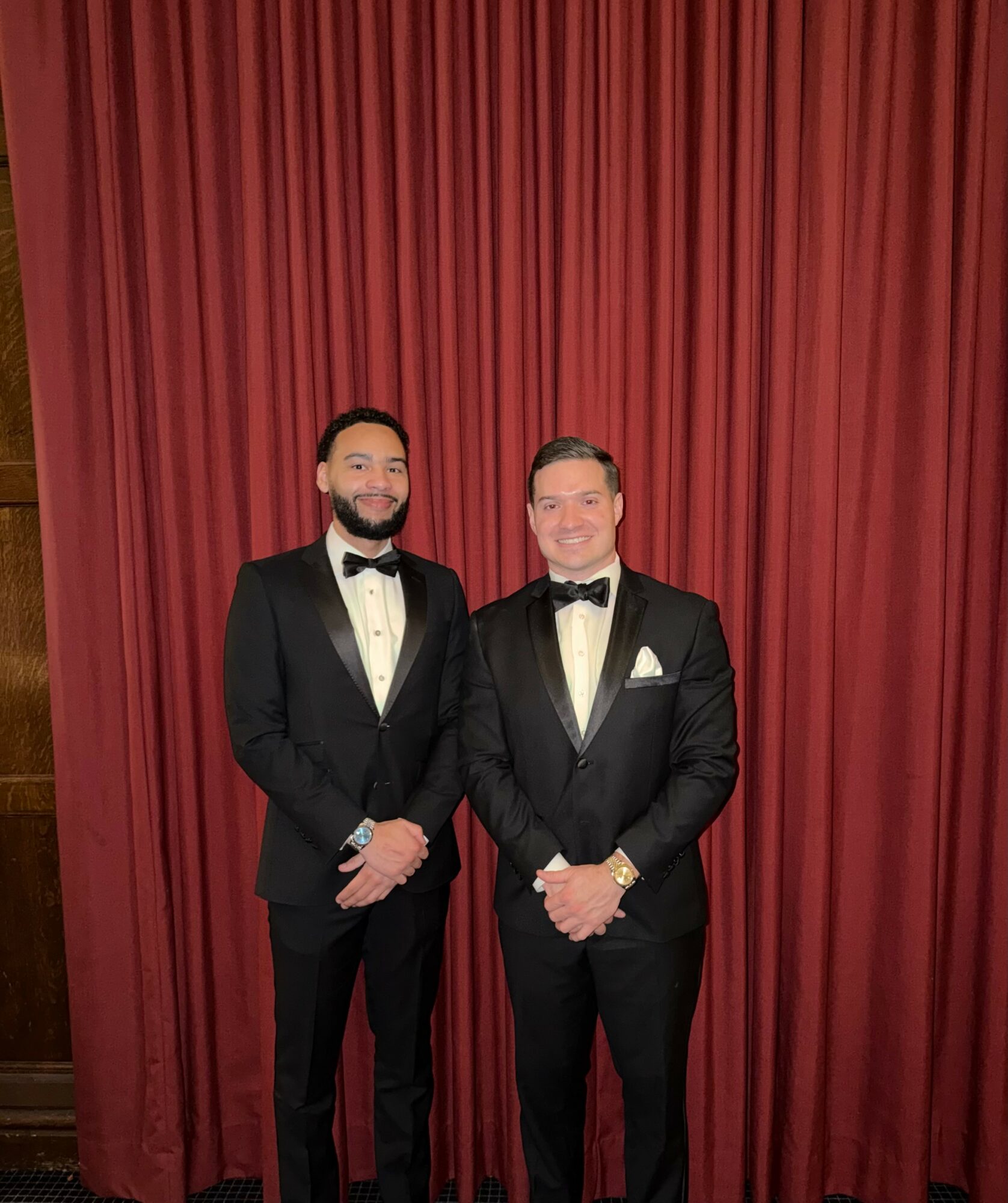 Two men in tuxedos stand in front of red curtains, smiling, with hands clasped in front, dressed formally for an event.