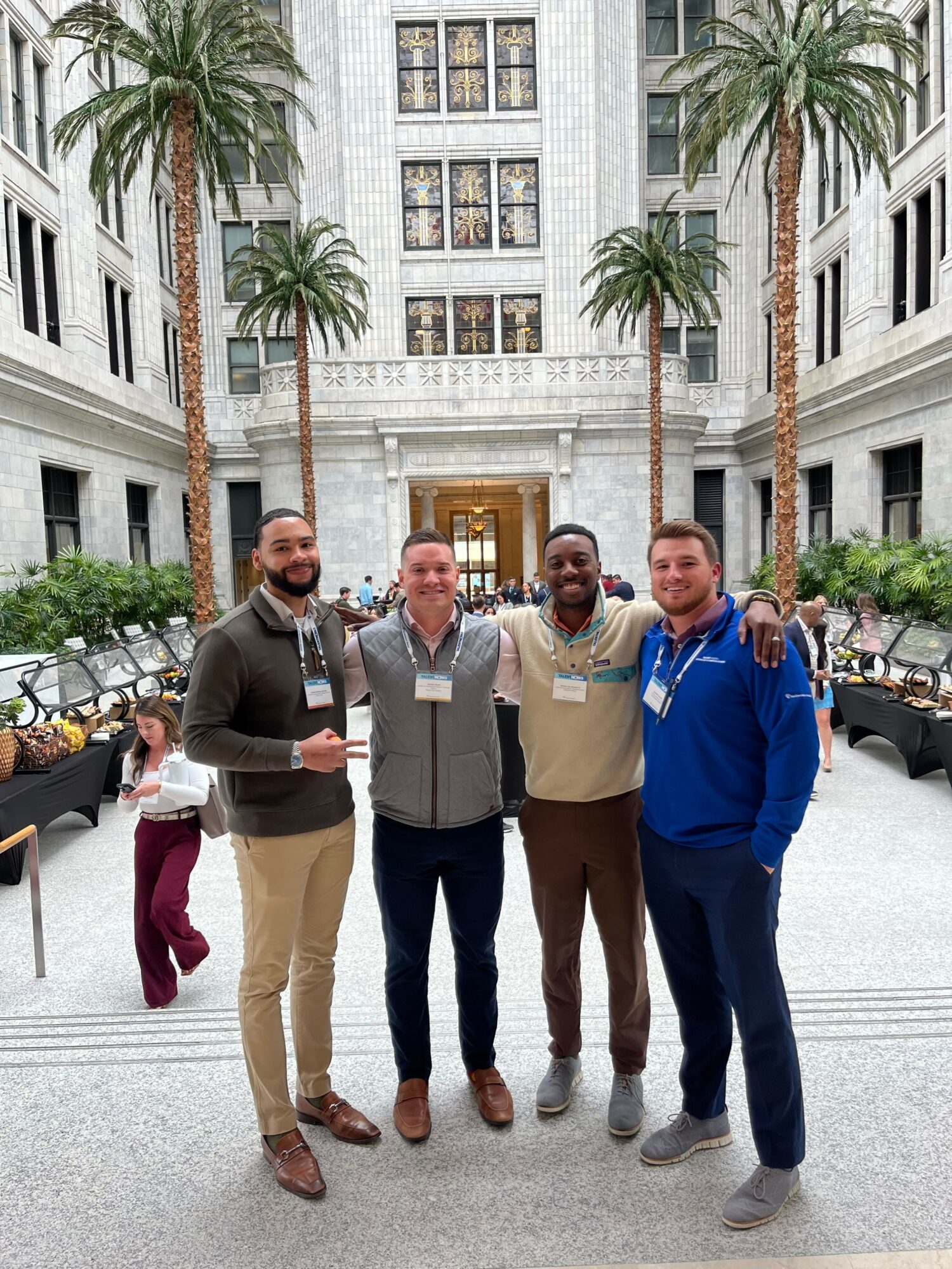 Four people standing outdoors in front of a building with palm trees, smiling and posing for a photo.