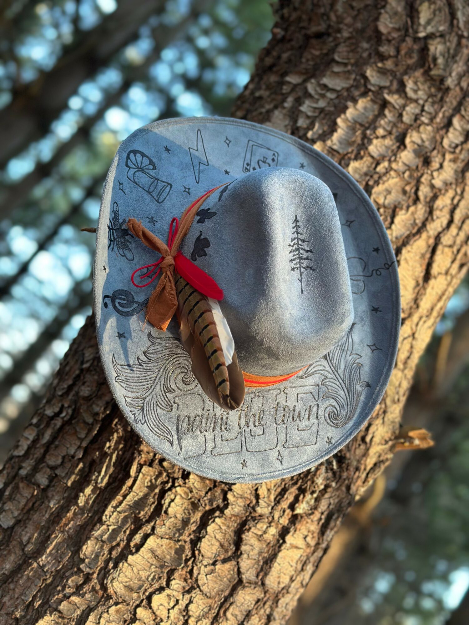 A silver hat with a feather and red ribbon is hanging on a tree trunk, with a background of blurred trees and sky.