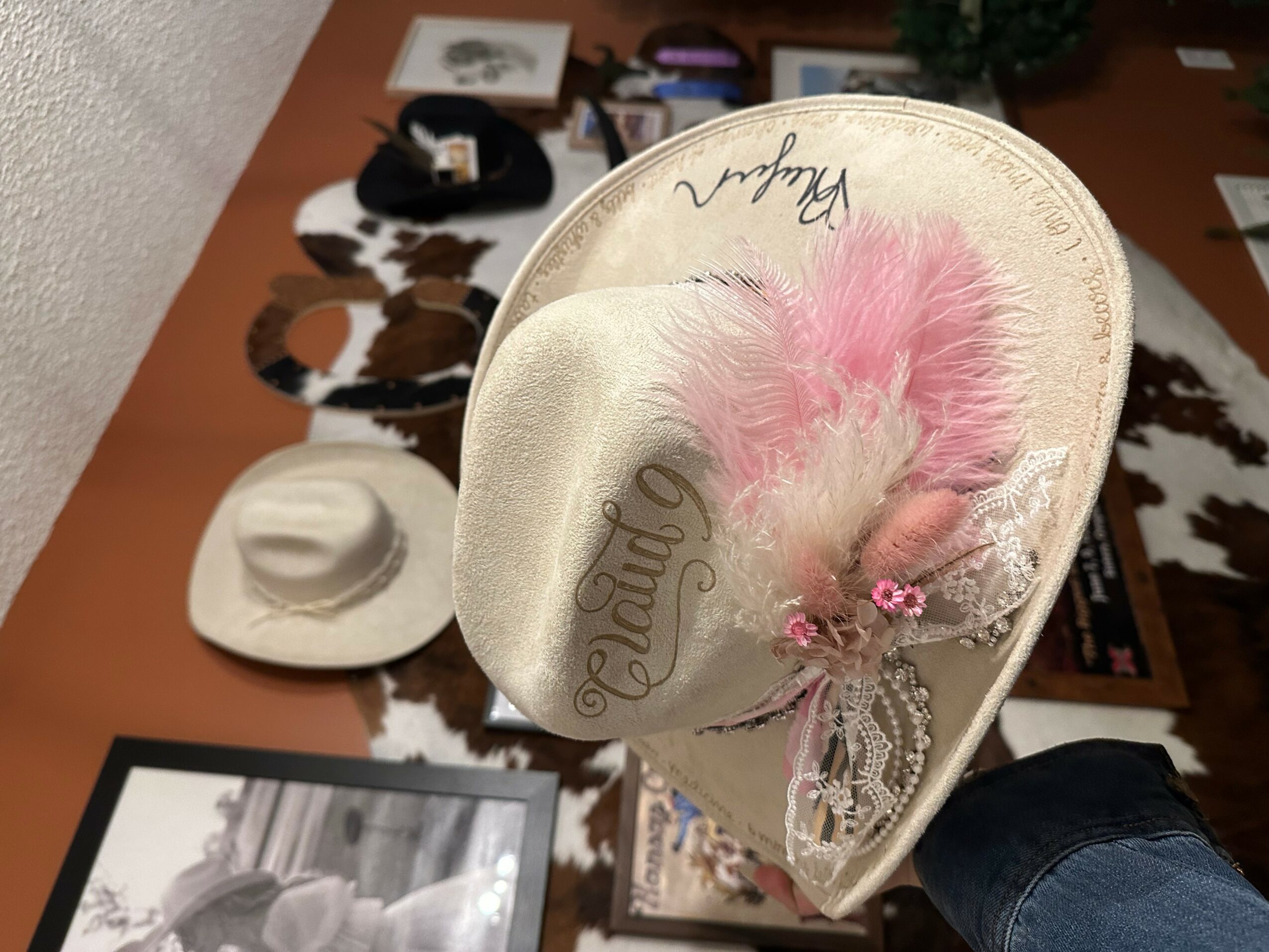 Person holding a white hat with pink feather and decorative flowers, other hats and items on table in background.