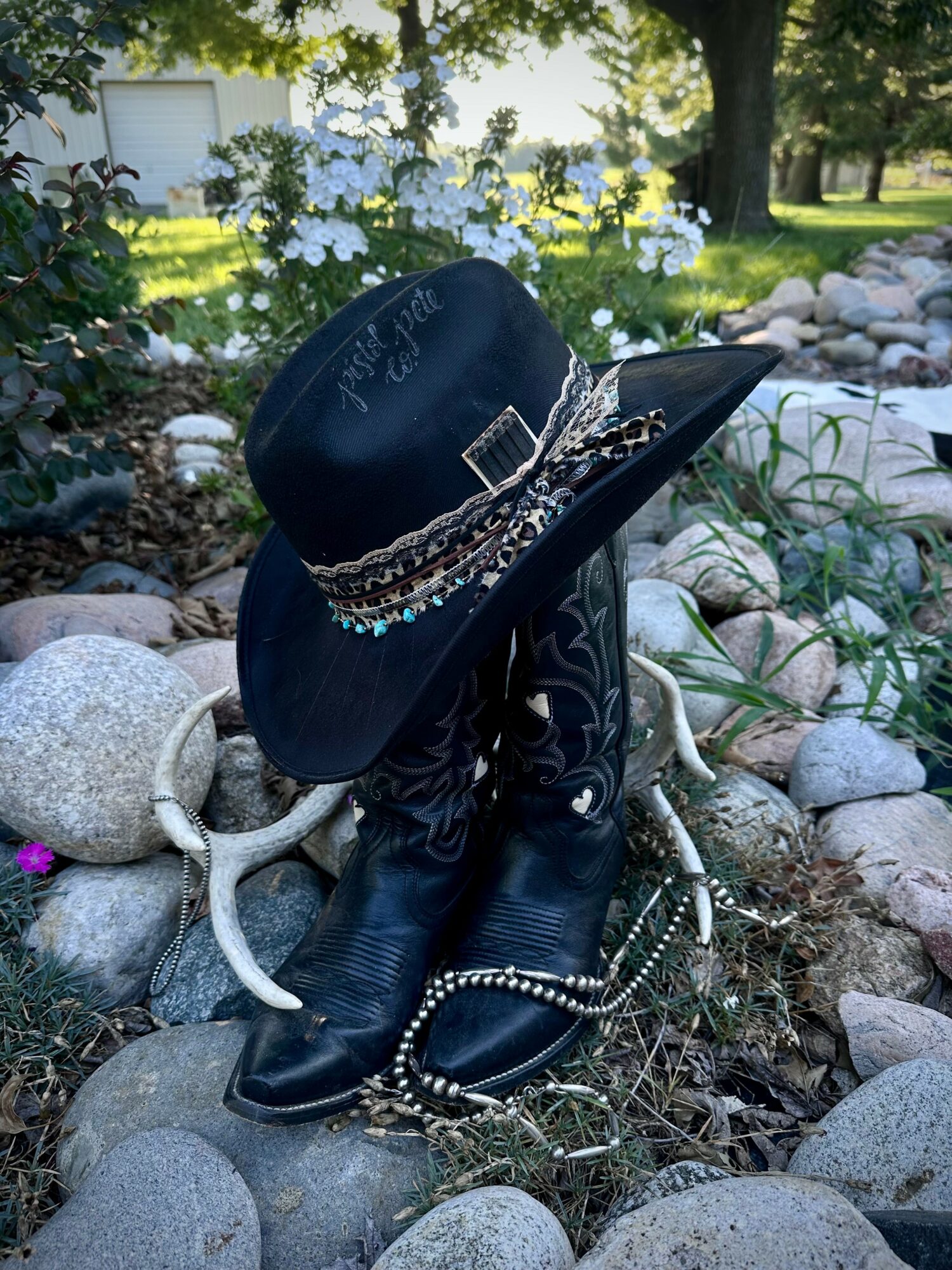 Cow skull with a black cowboy hat decorated with a band, surrounded by rocks and plants in a garden.