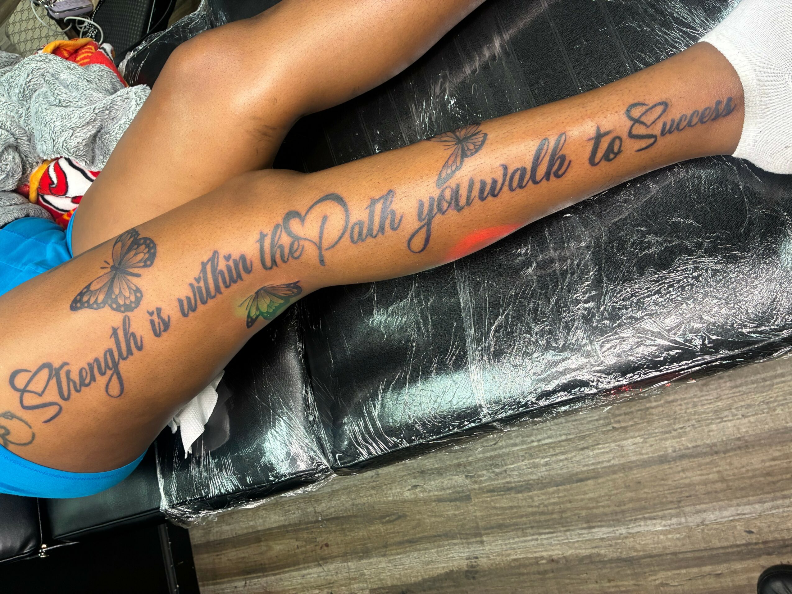 Tattooed arm with a butterfly and script text on a black surface, with a child's leg and shoes nearby.