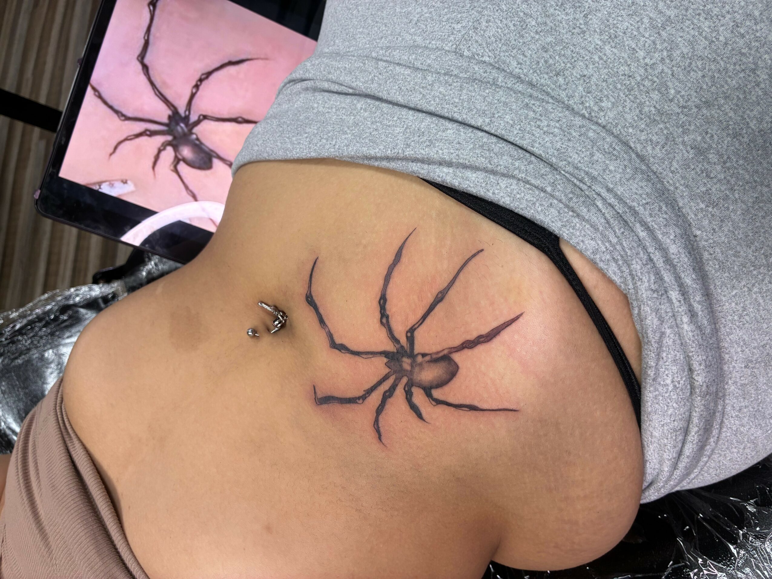 Person with a spider tattoo on their side, wearing a gray shirt and black underwear, lying down.