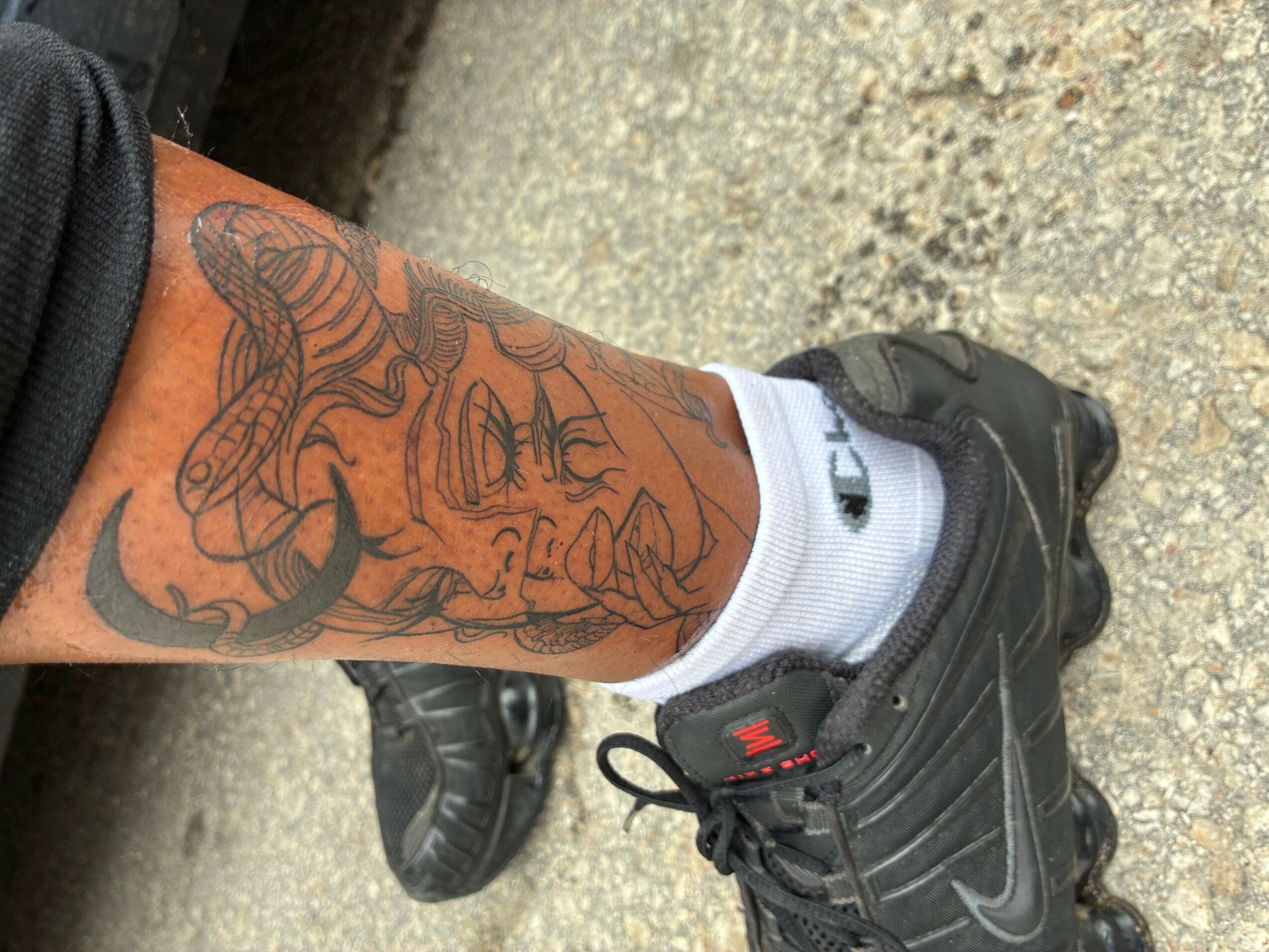 Person's leg with a tattoo of a woman's face, wearing a black sneaker and white sock, on a rough ground.