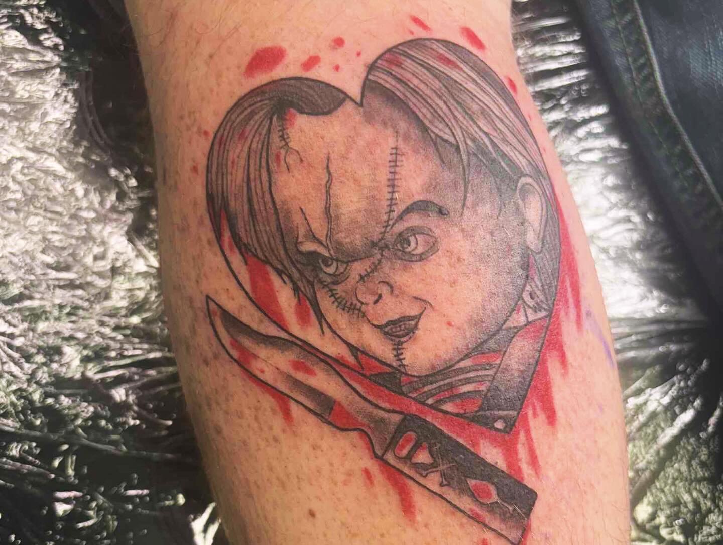 Tattoo of a child's face with a mischievous expression, holding a knife, surrounded by red splashes, on skin.