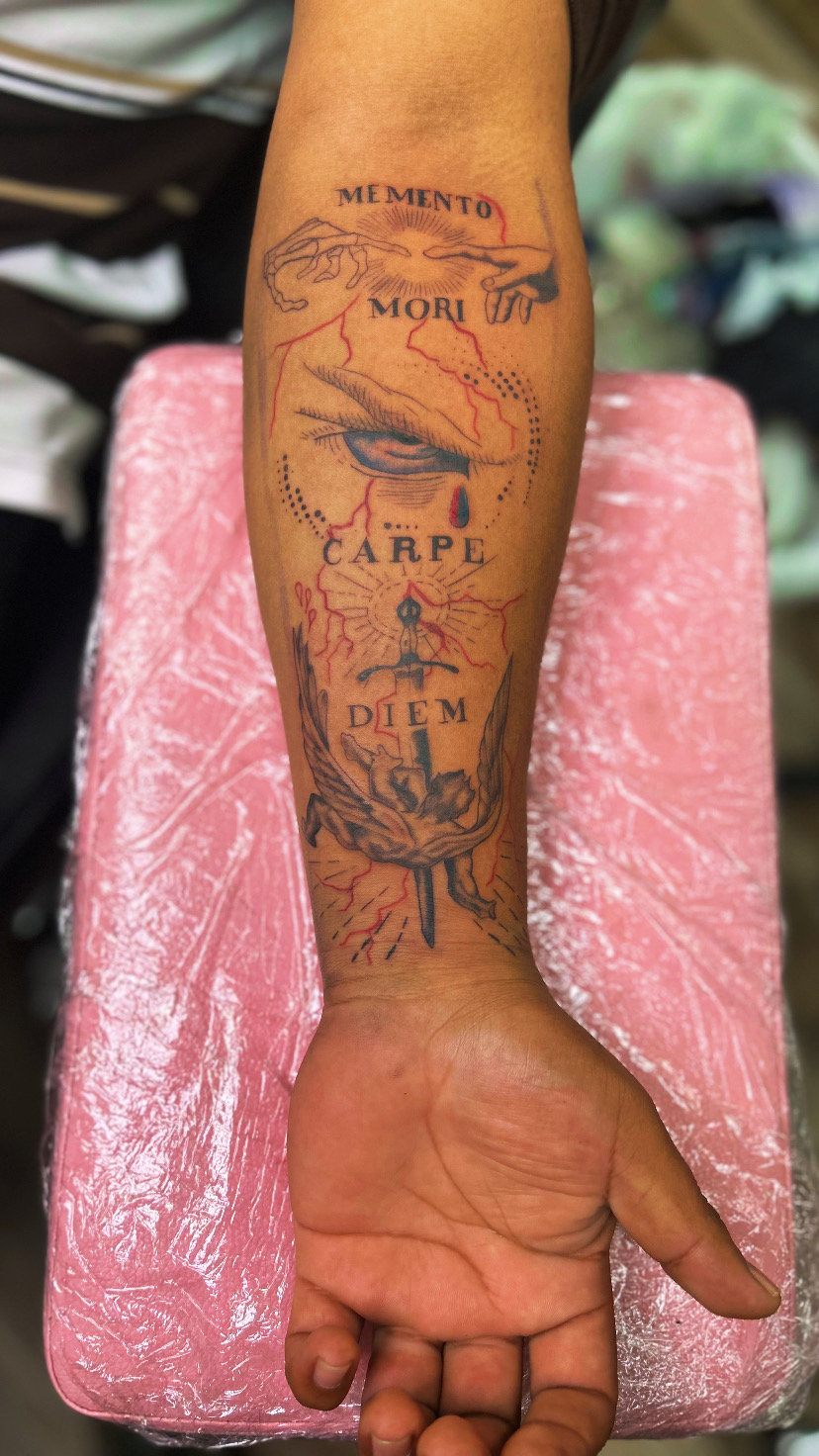 Forearm tattoo with Latin phrases, a hand, and decorative elements, resting on pink padding, with a blurred background.