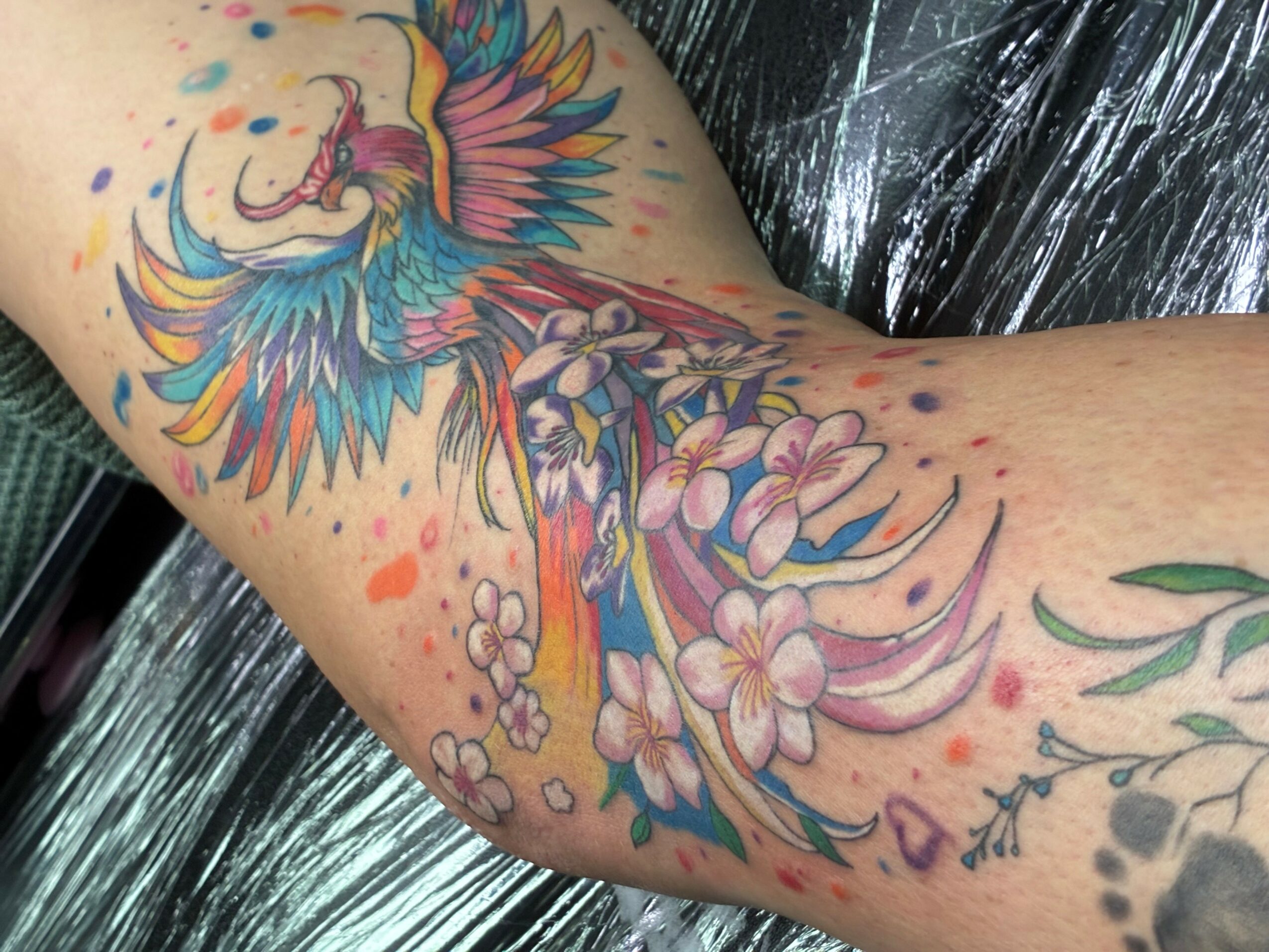 Colorful tattoo of a bird with wings, surrounded by flowers and abstract elements on a person's arm.