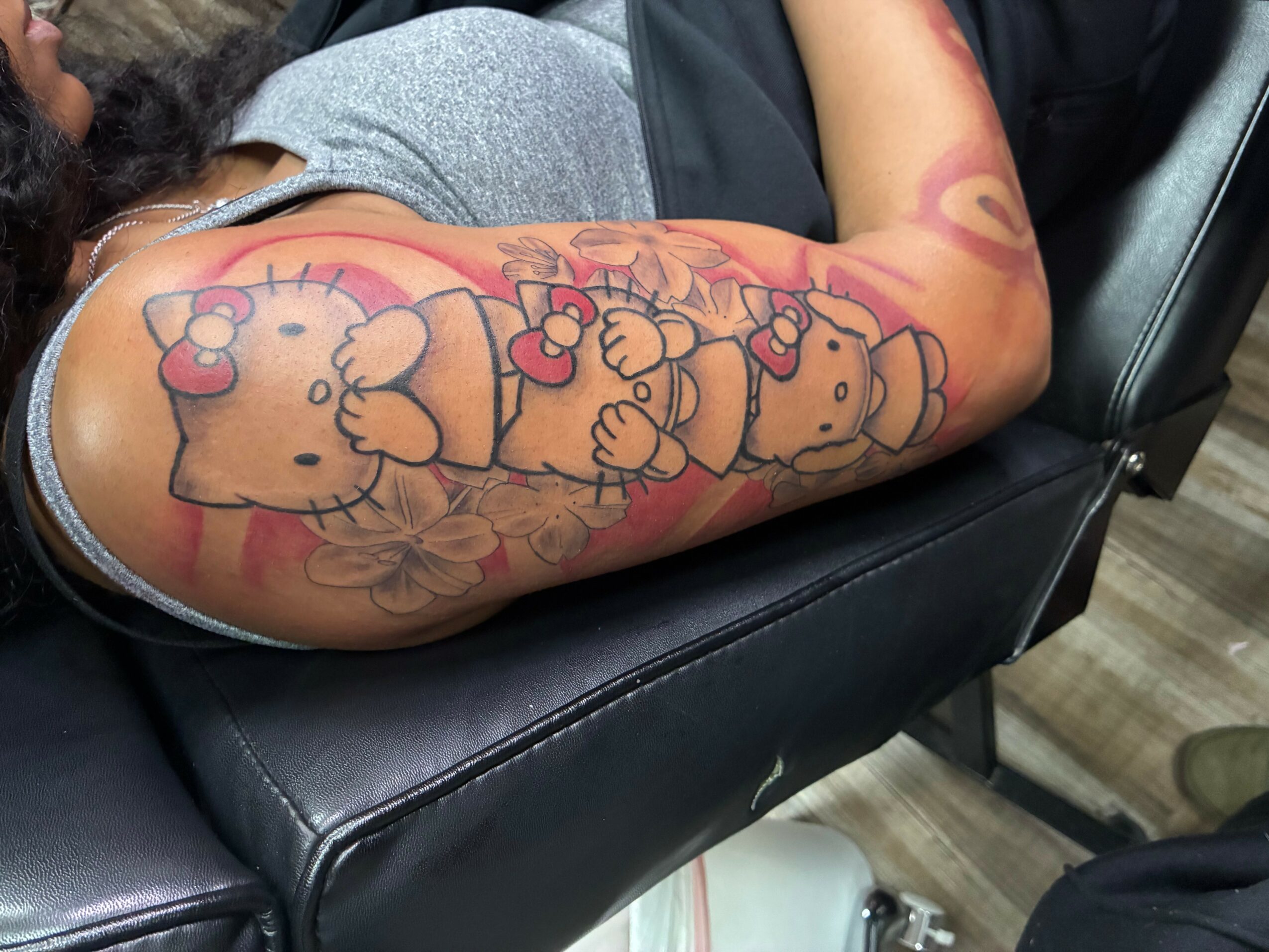 Tattoo of cartoon bears holding hearts on person's arm, with pink and red colors, resting on black leather surface.