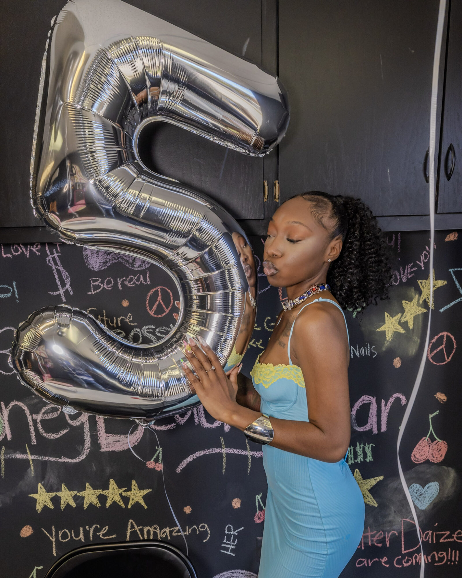 Young woman in a blue dress holding a large silver balloon shaped as the number five, standing in front of a colorful chalkboard wall.