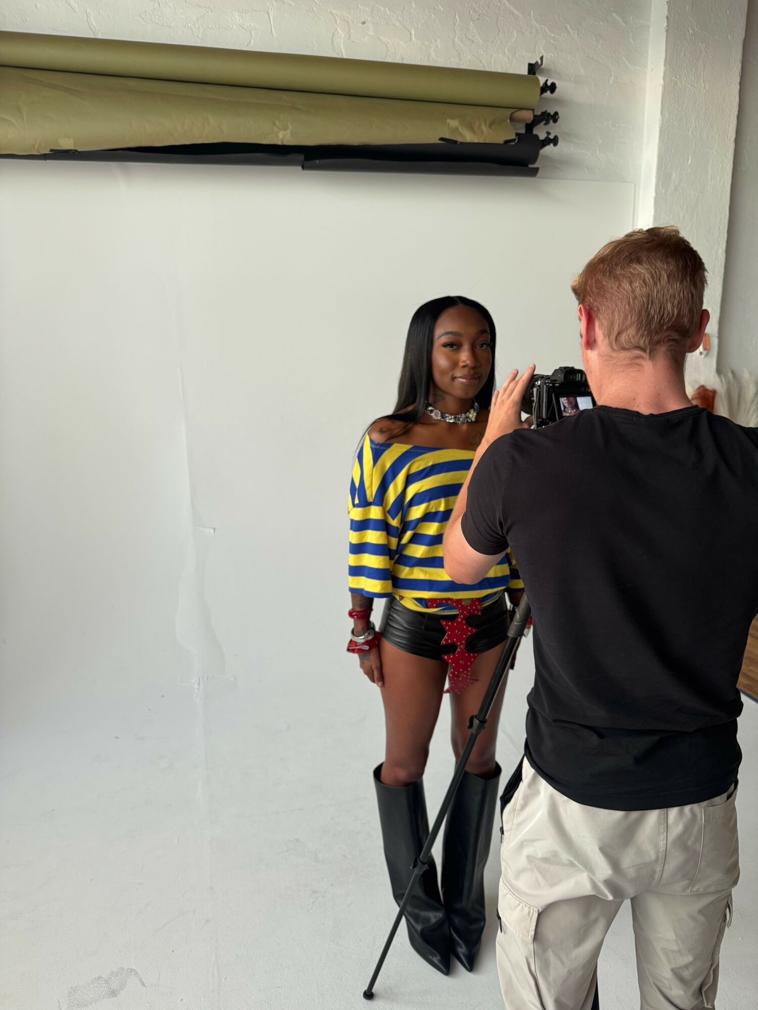 Person taking a photo of a woman standing against a white wall, woman wearing a striped shirt and shorts, man in black shirt and light pants holding a camera, indoor setting.
