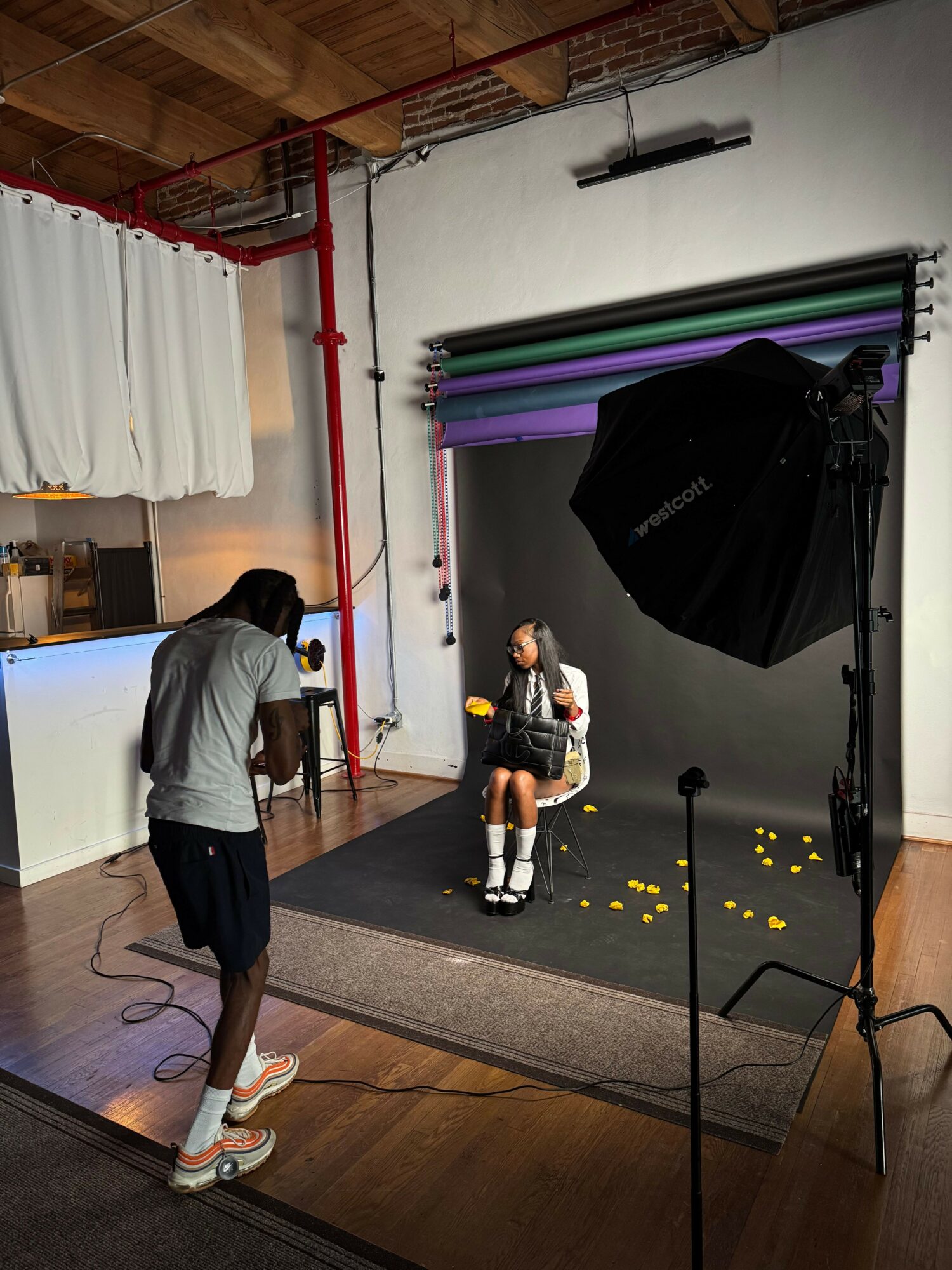 Person standing in a photography studio with a black backdrop, camera, and lighting equipment, while another person adjusts a tripod.