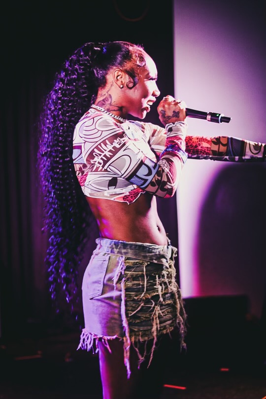 Performer with long curly hair singing into a microphone on stage, wearing a patterned crop top and ripped shorts.