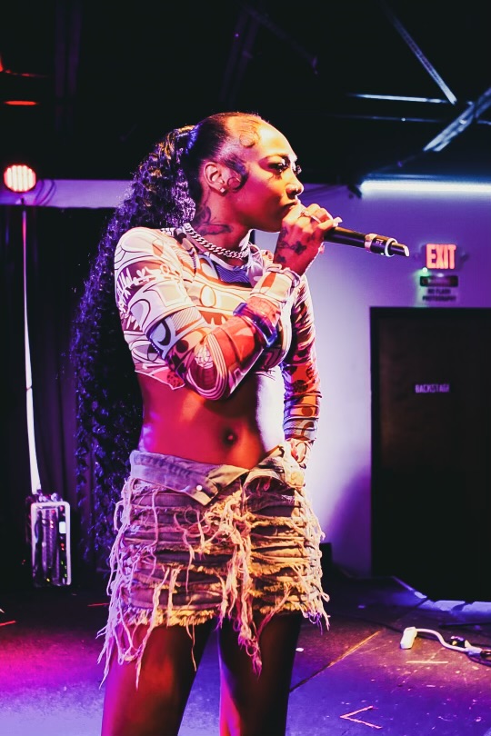 Performer singing into a microphone on stage with purple and blue lighting, wearing a patterned top and ripped shorts.