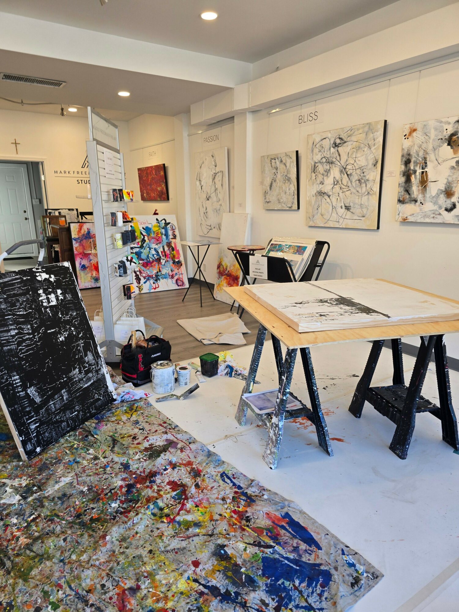Art gallery with paintings on walls, a table with art supplies, and a colorful, paint-splattered floor.