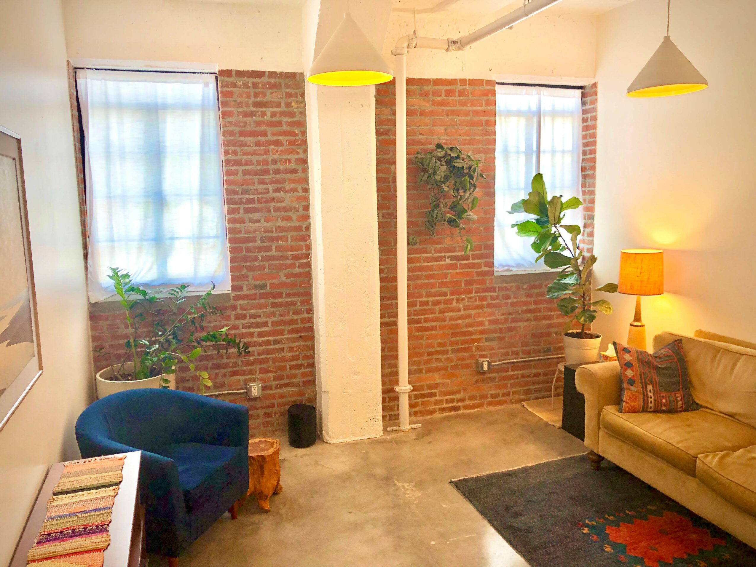Living room with brick wall, two windows with curtains, a blue armchair, a beige sofa, a lamp, and potted plants.