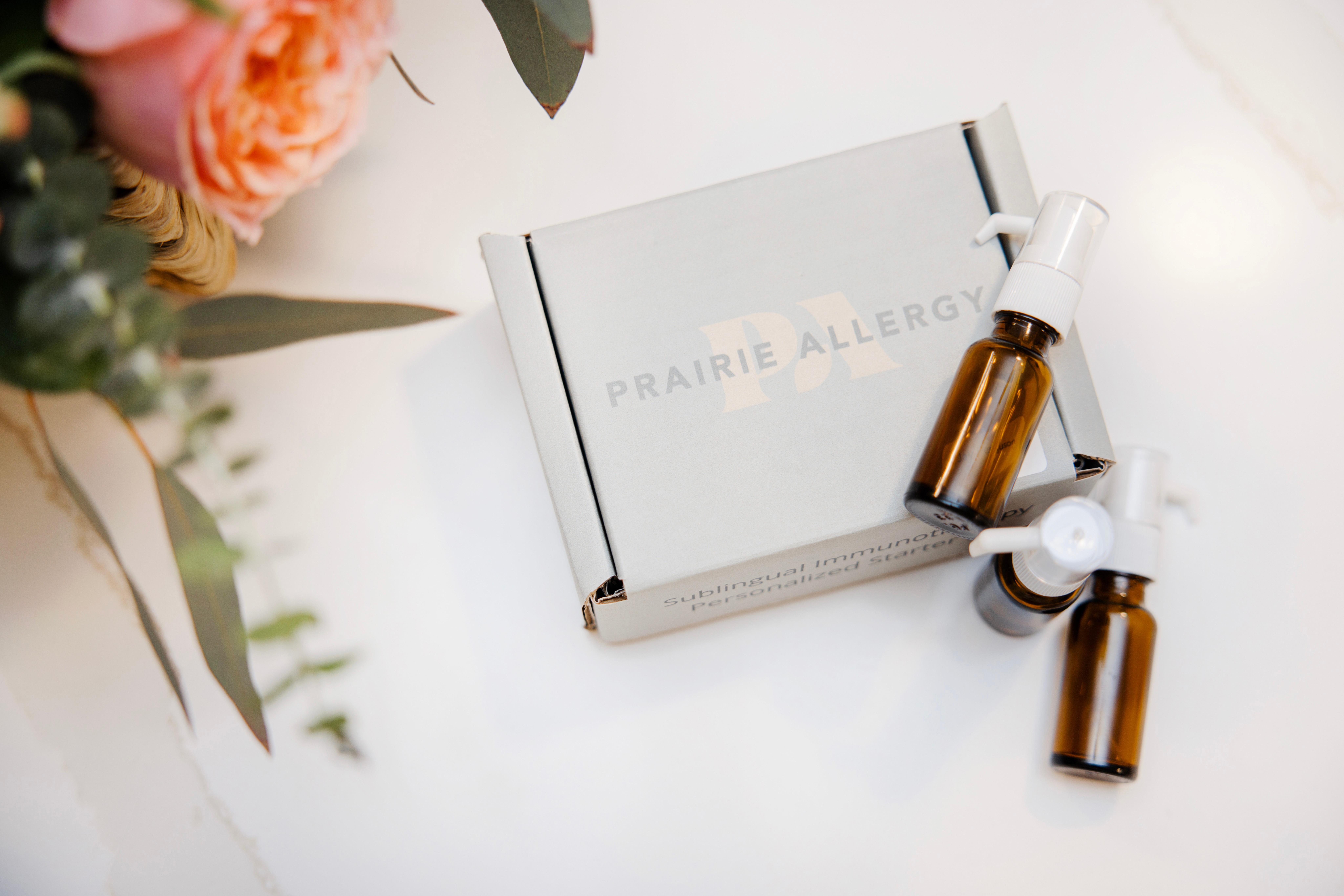 Box labeled Prairie Allergy with two small amber glass bottles and white dropper caps on a white surface, partially visible flowers on the left