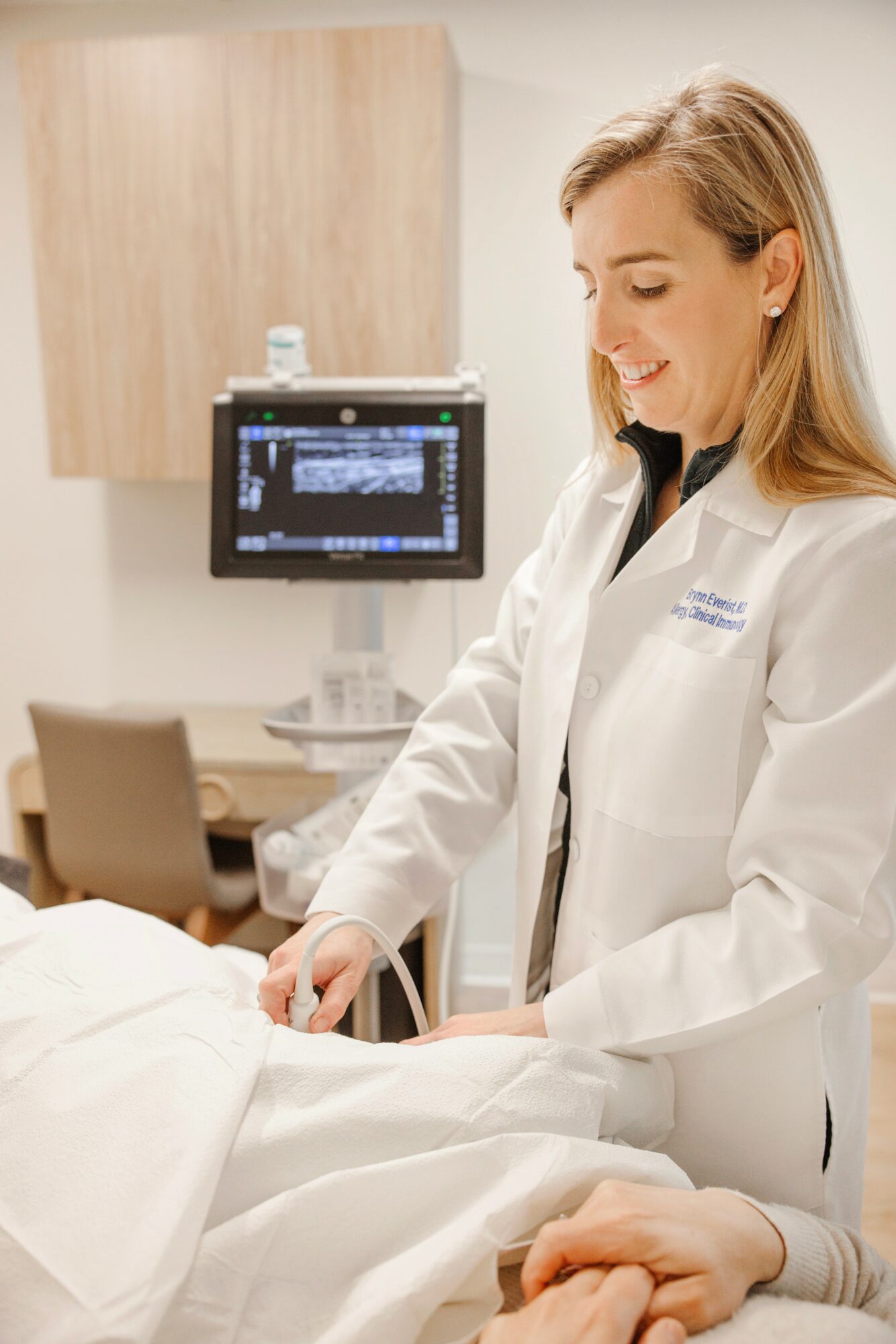 Female healthcare professional smiling, wearing a white coat, attending to a patient in a medical office.
