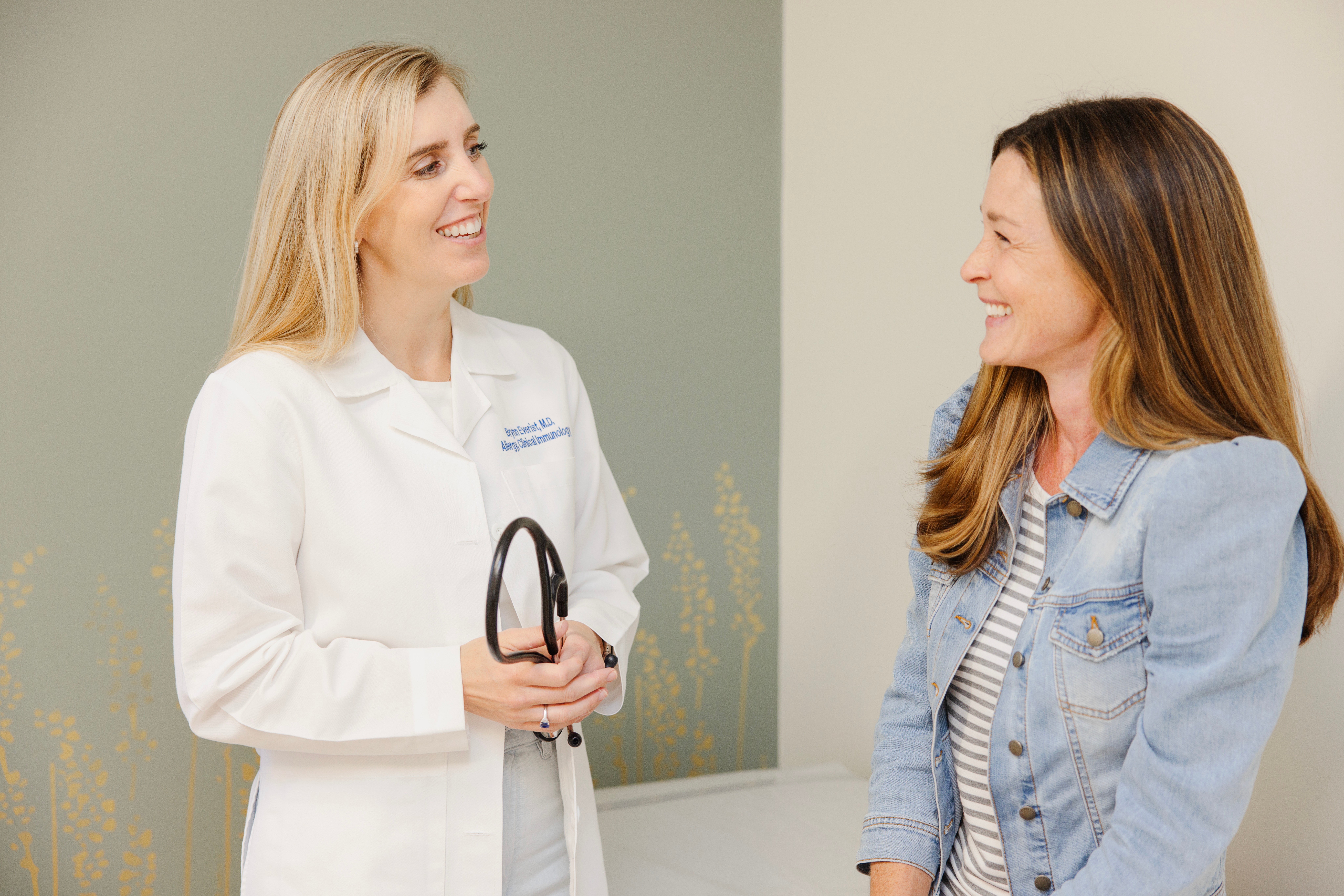 Two women smiling and talking indoors, one in a white coat holding a stethoscope, the other in a denim jacket.