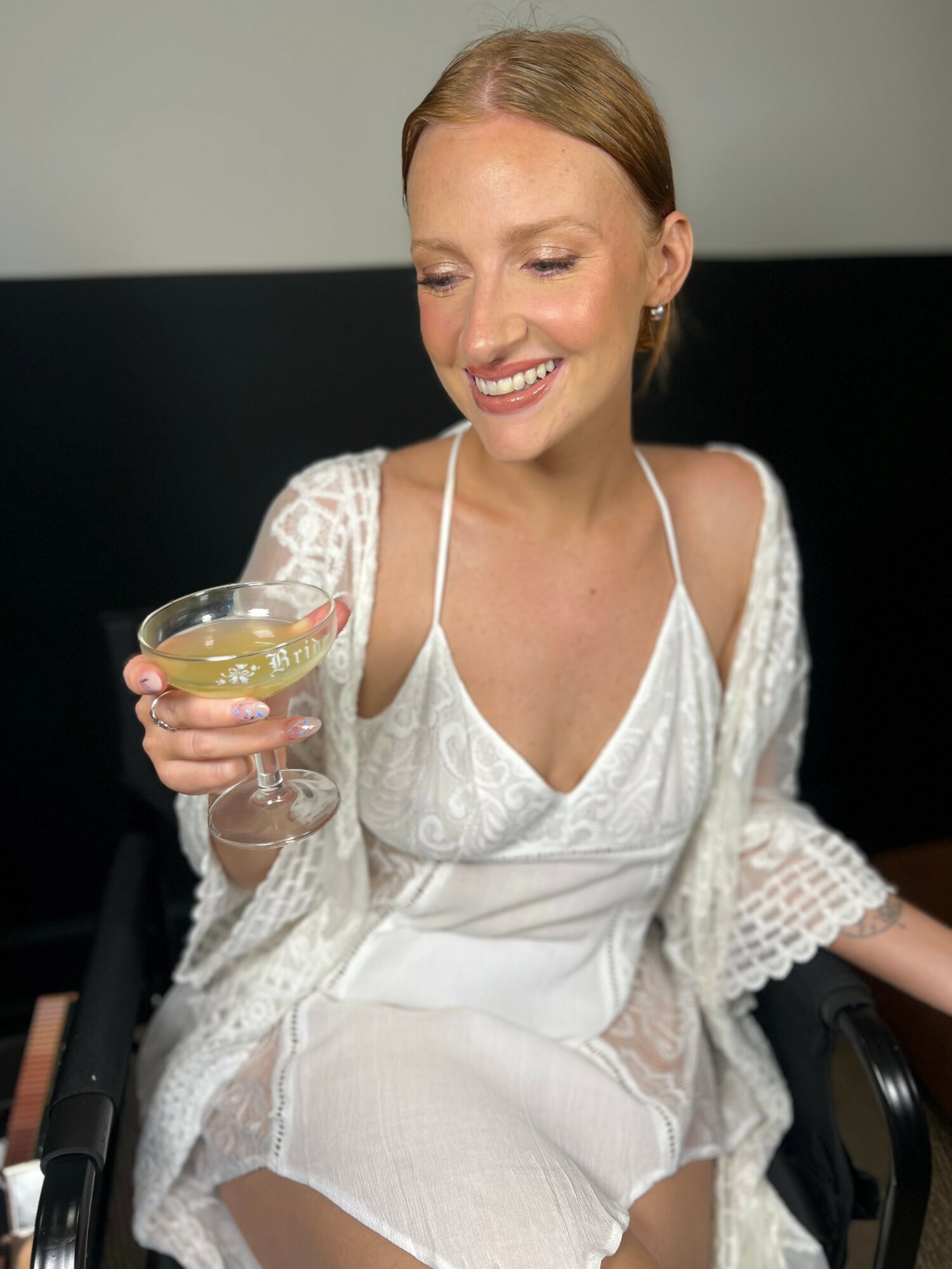 Smiling woman with red hair in a white dress holding a cocktail glass, sitting in a chair.
