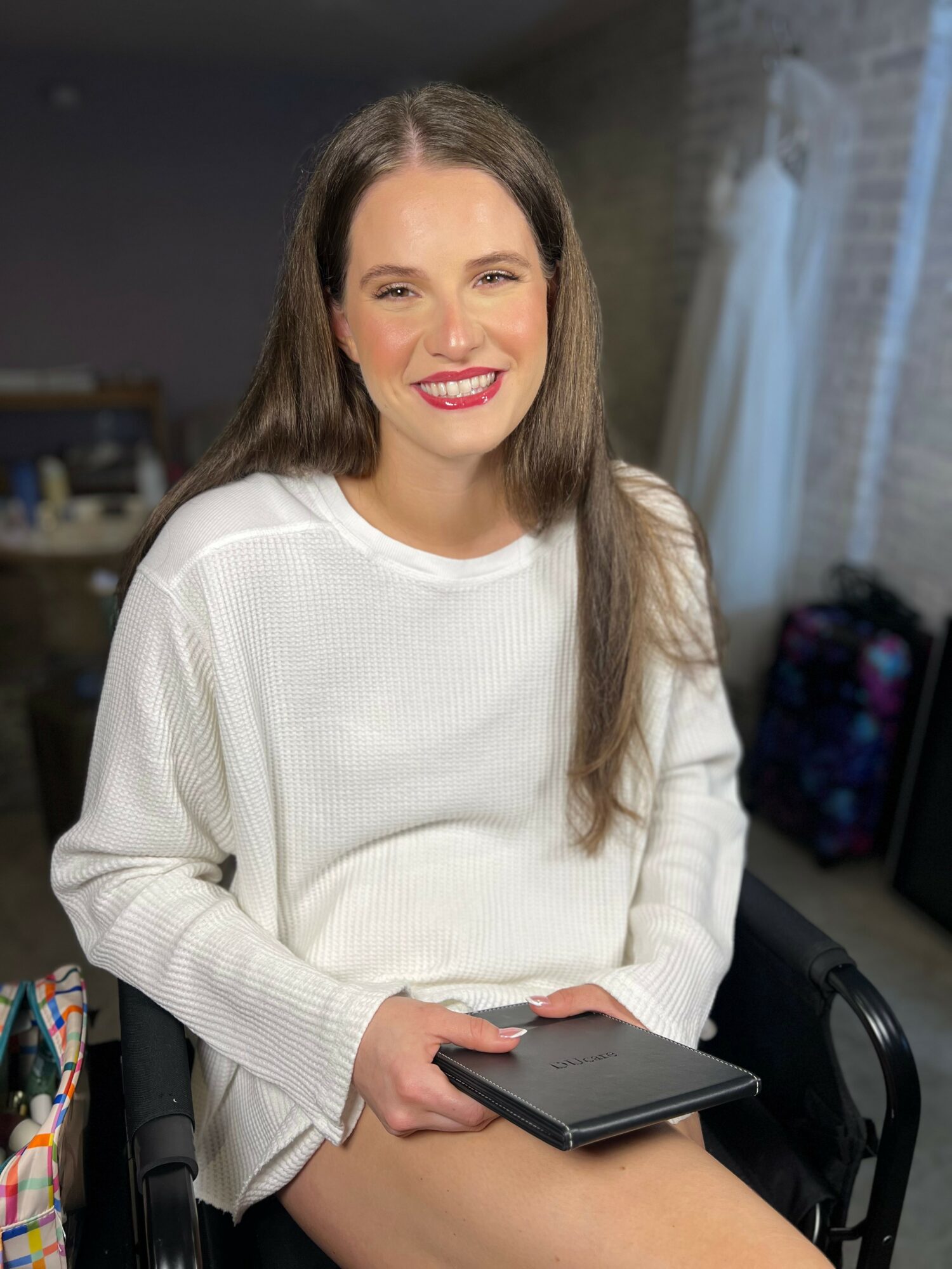 Smiling woman with long brown hair in a white sweater holding a tablet, sitting indoors near a window.
