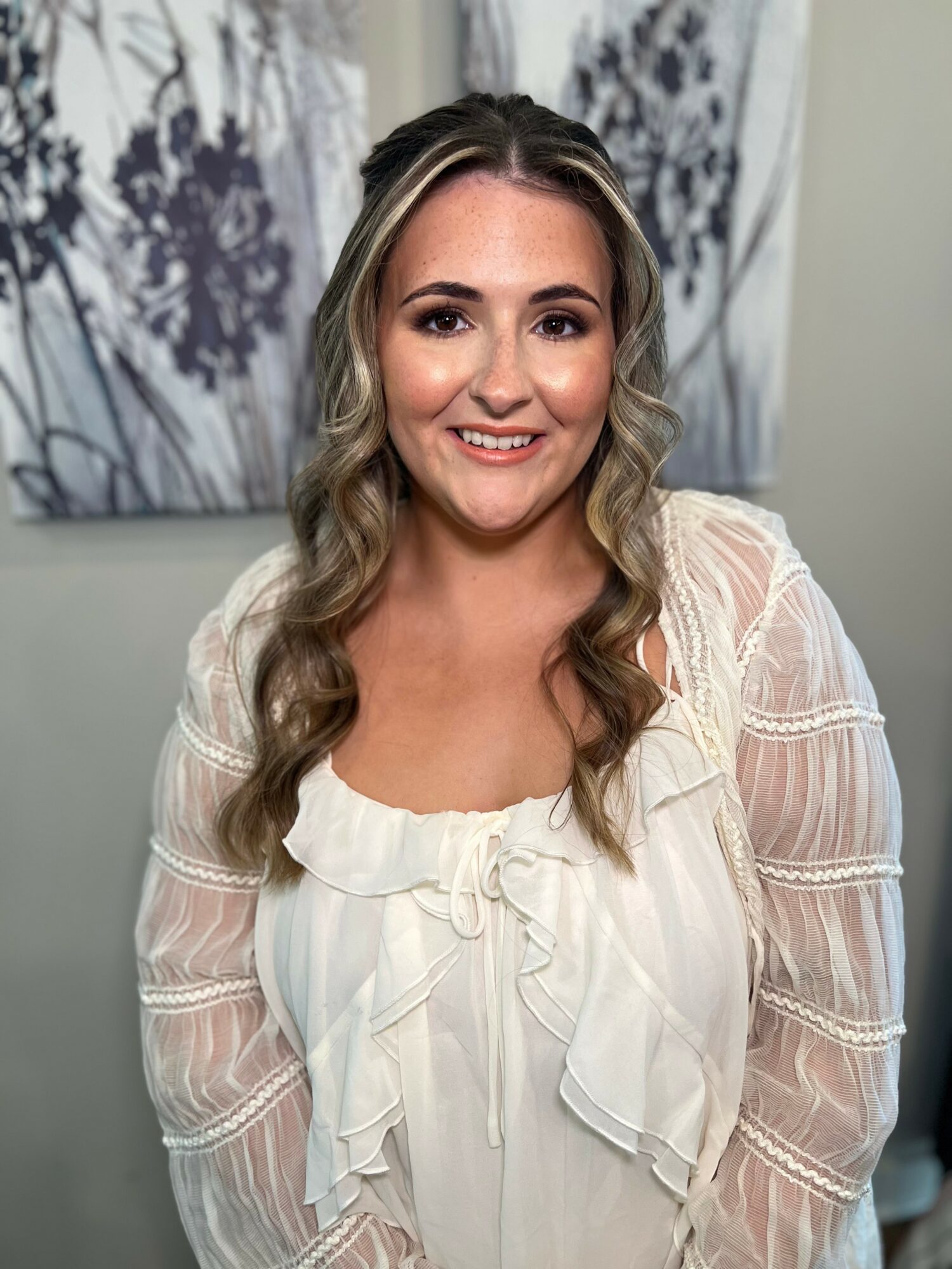 Young woman with long wavy hair smiling, wearing a white blouse with ruffles, standing indoors in front of a floral artwork.