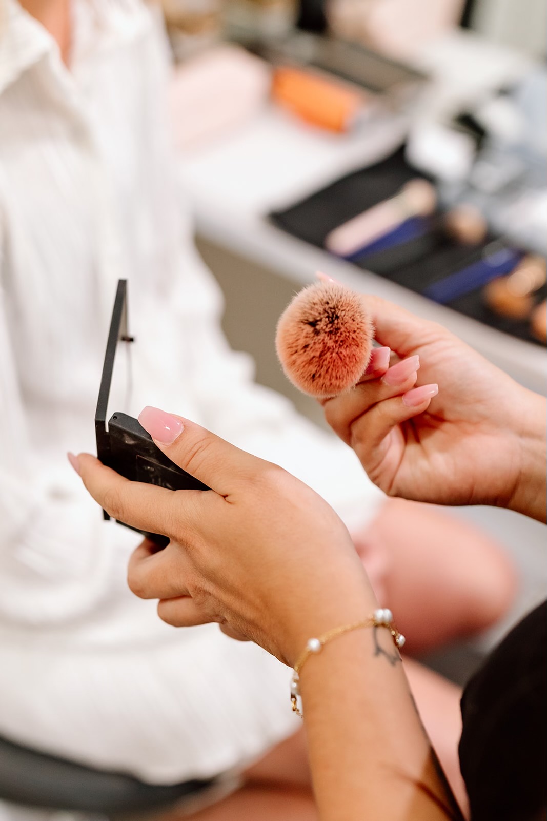 Person holding a makeup brush and a compact mirror, with makeup tools on a table in the background.