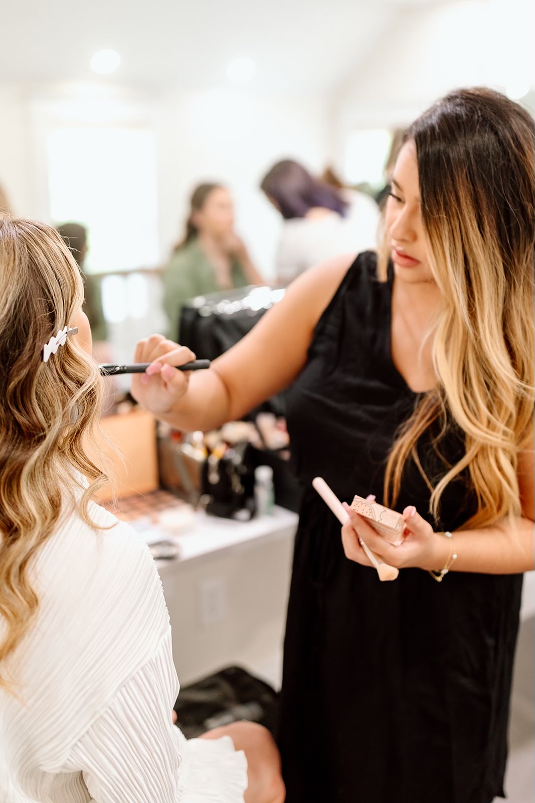 Makeup artist applying makeup to a woman in a white top, with a blurred background of people and makeup products.