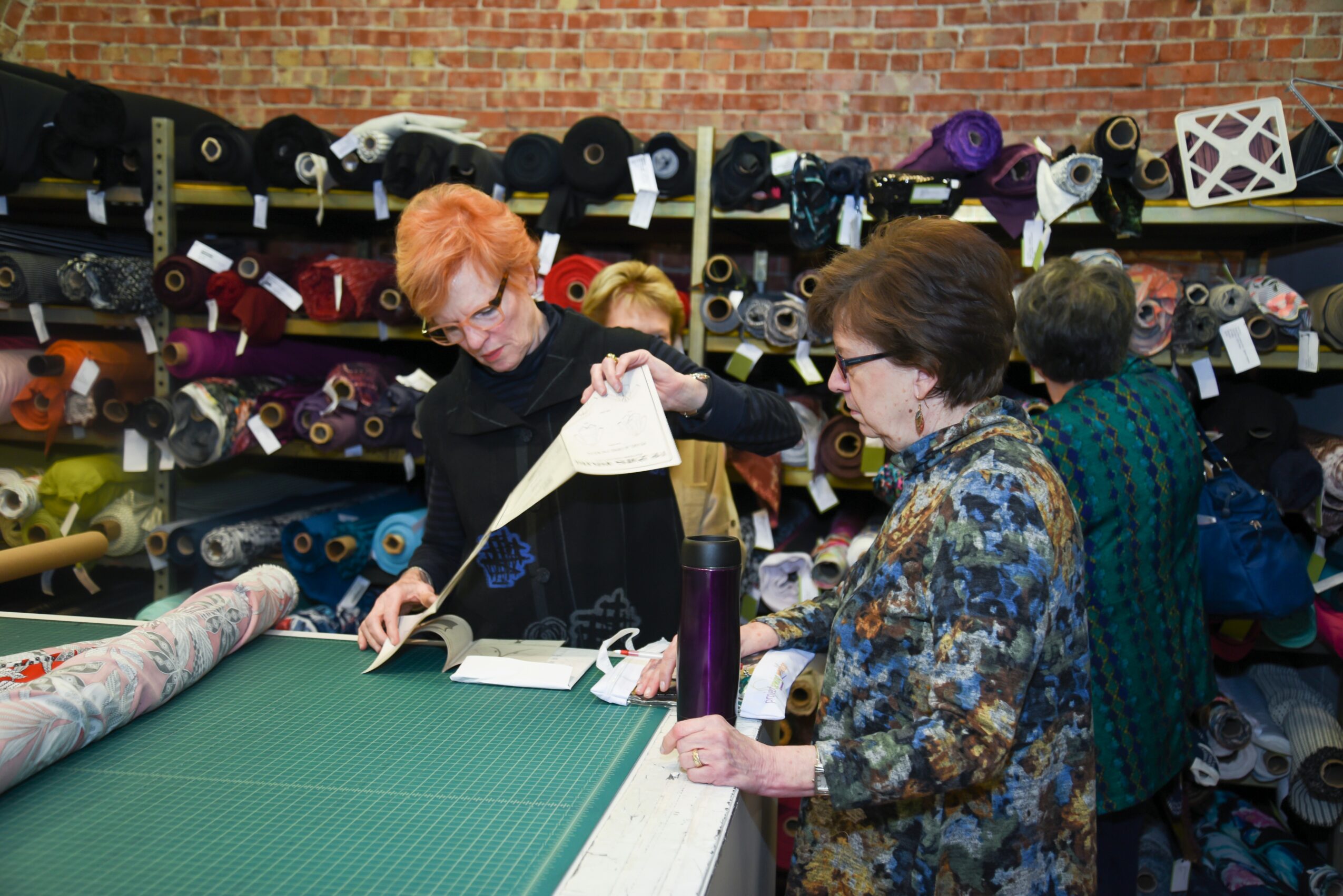 Group of people in a fabric store looking at a book, shelves filled with yarn and fabric rolls in the background.